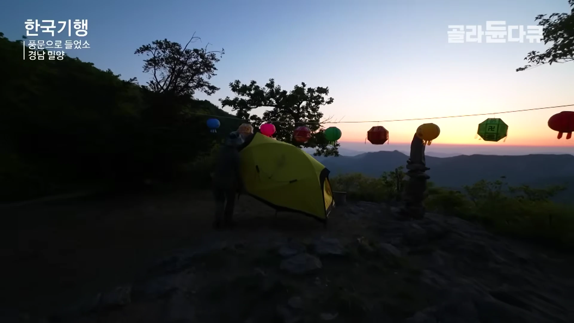 노을이 지는 산 정상에서 한 사람이 산장 근처에 노란색 텐트를 설치하고 있으며, 줄에 매달린 연등들이 저녁 하늘 아래 나란히 걸려 있다.