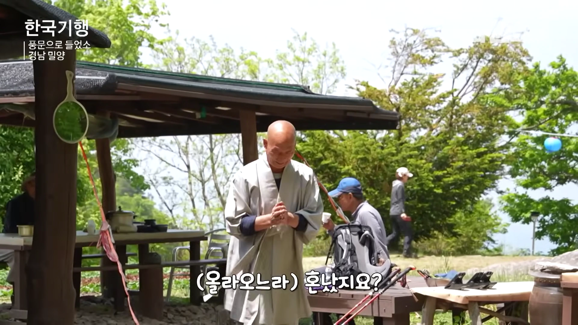 머리를 깎은 승려가 야외 암자 마당에서 두 손을 모아 합장하며 인사를 건네고 있고, 그 뒤로 등산객들이 쉬고 있는 모습