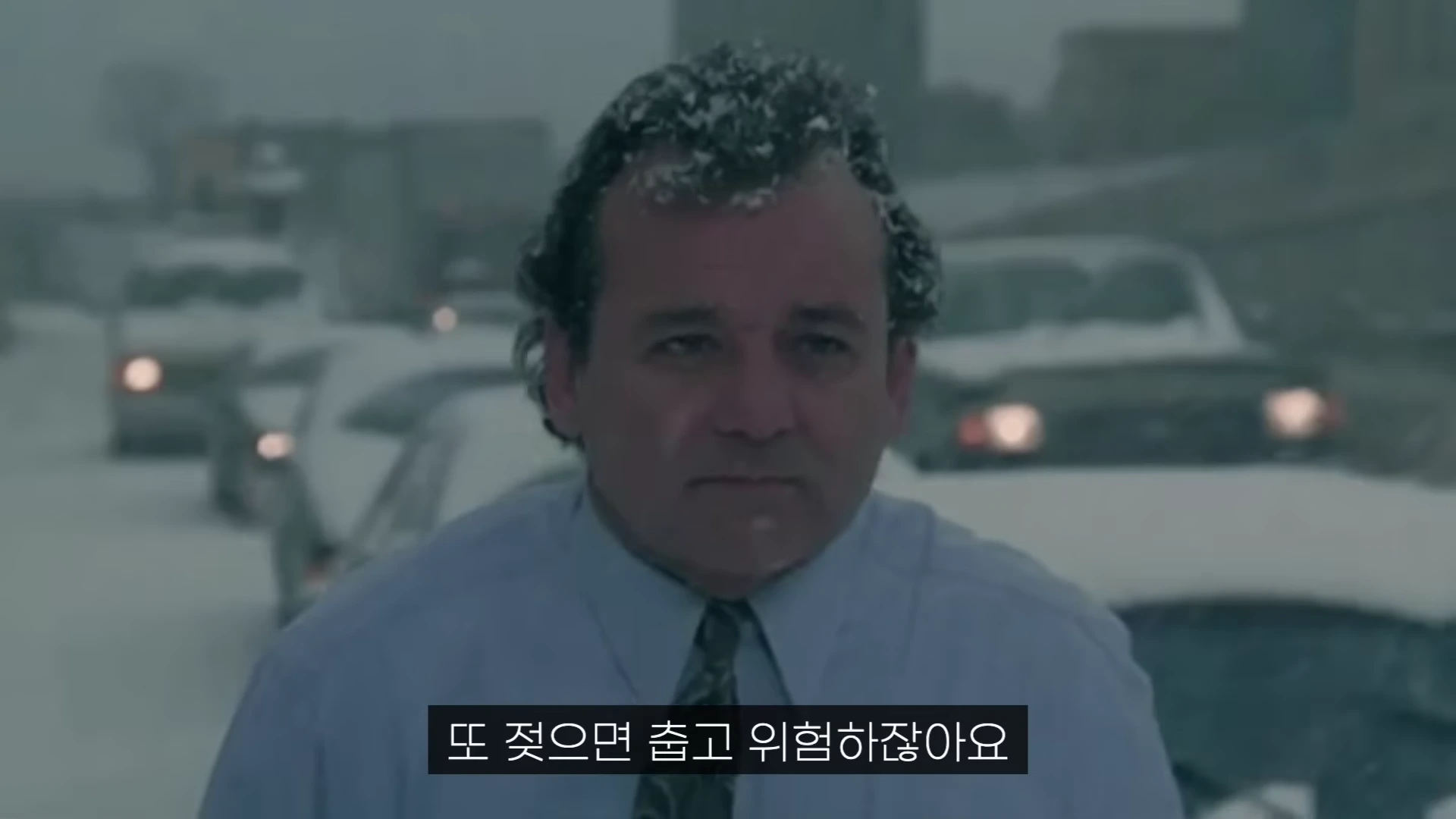 눈이 내리는 거리에서 셔츠와 넥타이 차림의 한 남성이 머리에 눈을 맞으며 정면을 응시하고 있는 영화 장면이다.