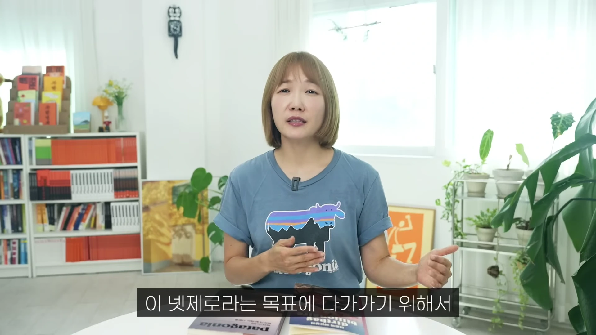단발머리를 한 여성이 밝은 실내에서 파타고니아 로고가 그려진 파란색 티셔츠를 입고 이야기를 나누고 있으며, 앞에는 파타고니아 관련 책들이 놓여 있다.