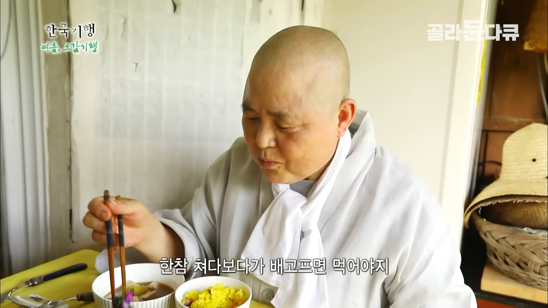 회색 승복을 입은 스님이 식탁 앞에서 젓가락으로 음식을 집어 들고 있는 모습이며 화면 하단에 자막이 있다.