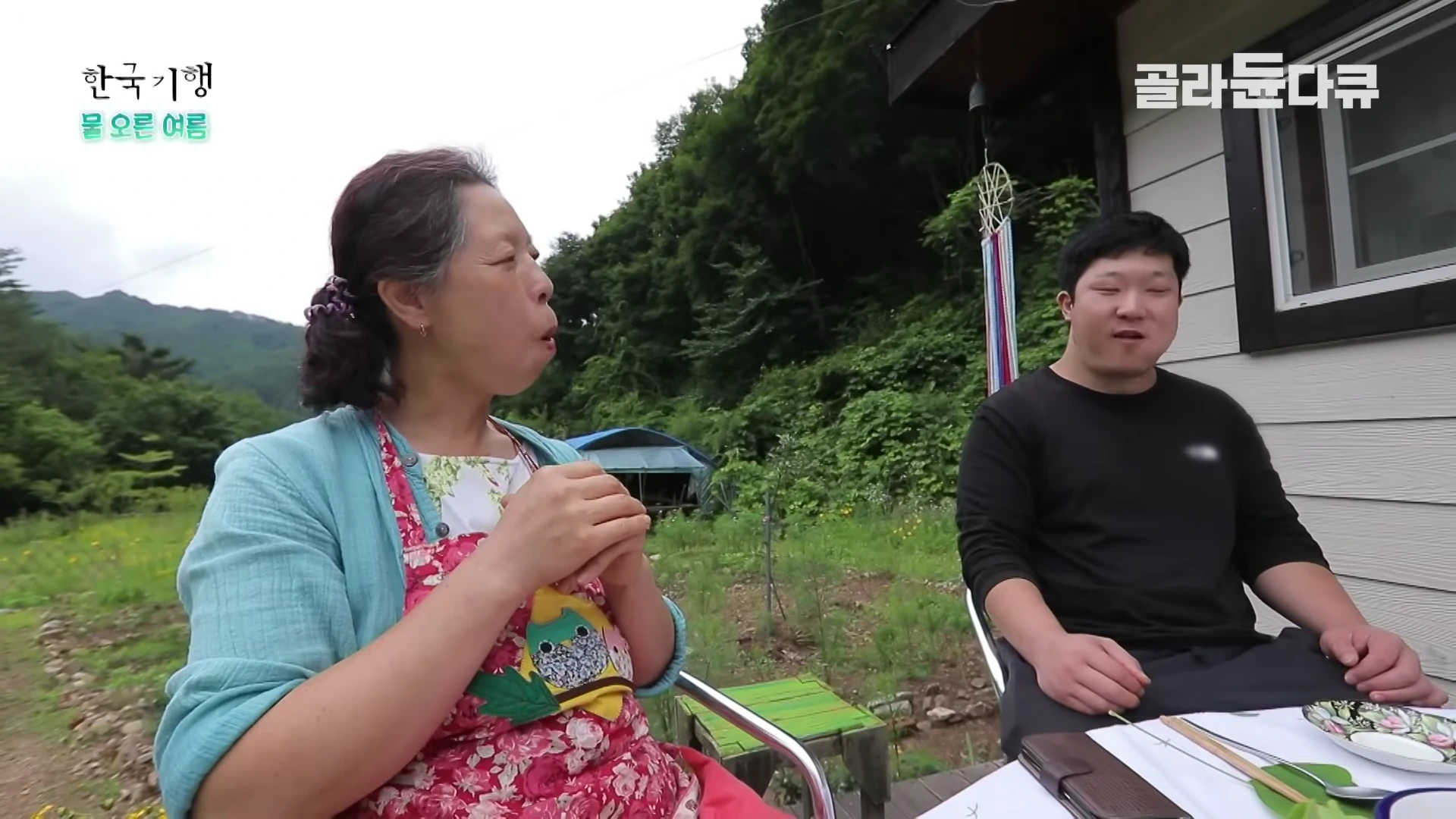 숲속 오두막 앞 야외 테이블에 마주 앉은 중년 여성과 젊은 남성이 함께 음식을 먹으며 이야기를 나누고 있다.