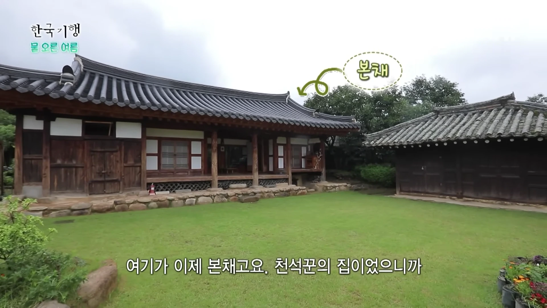 푸른 잔디 마당 뒤로 기와지붕을 얹은 낡은 한옥 본채와 부속 건물이 배치되어 있고, 본채 위로 '본채'라는 글자와 화살표가 표시된 화면.