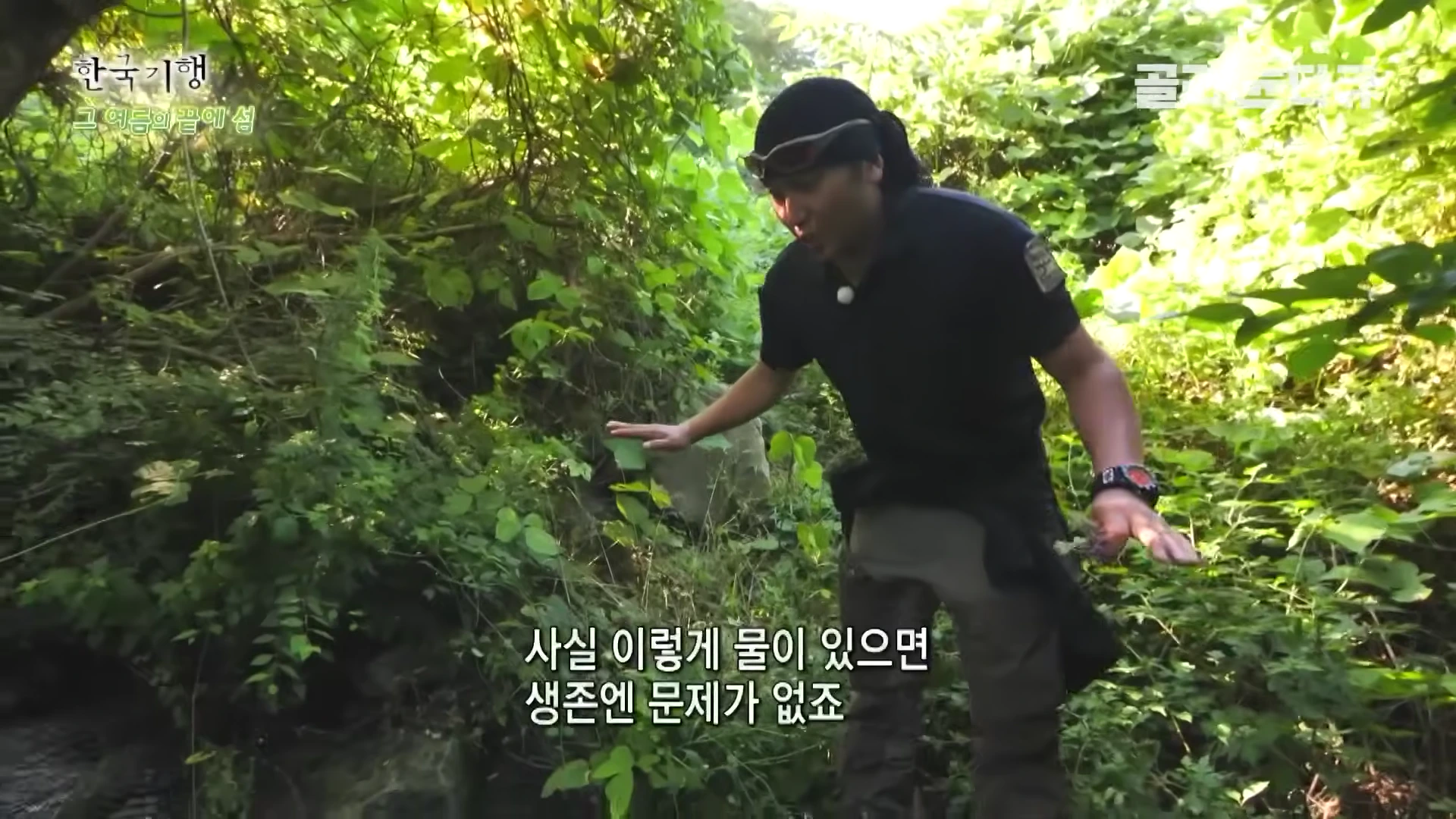 검은색 복장을 입은 남성이 우거진 숲속 계곡 근처에서 손으로 주변 지형을 가리키며 설명하고 있는 모습