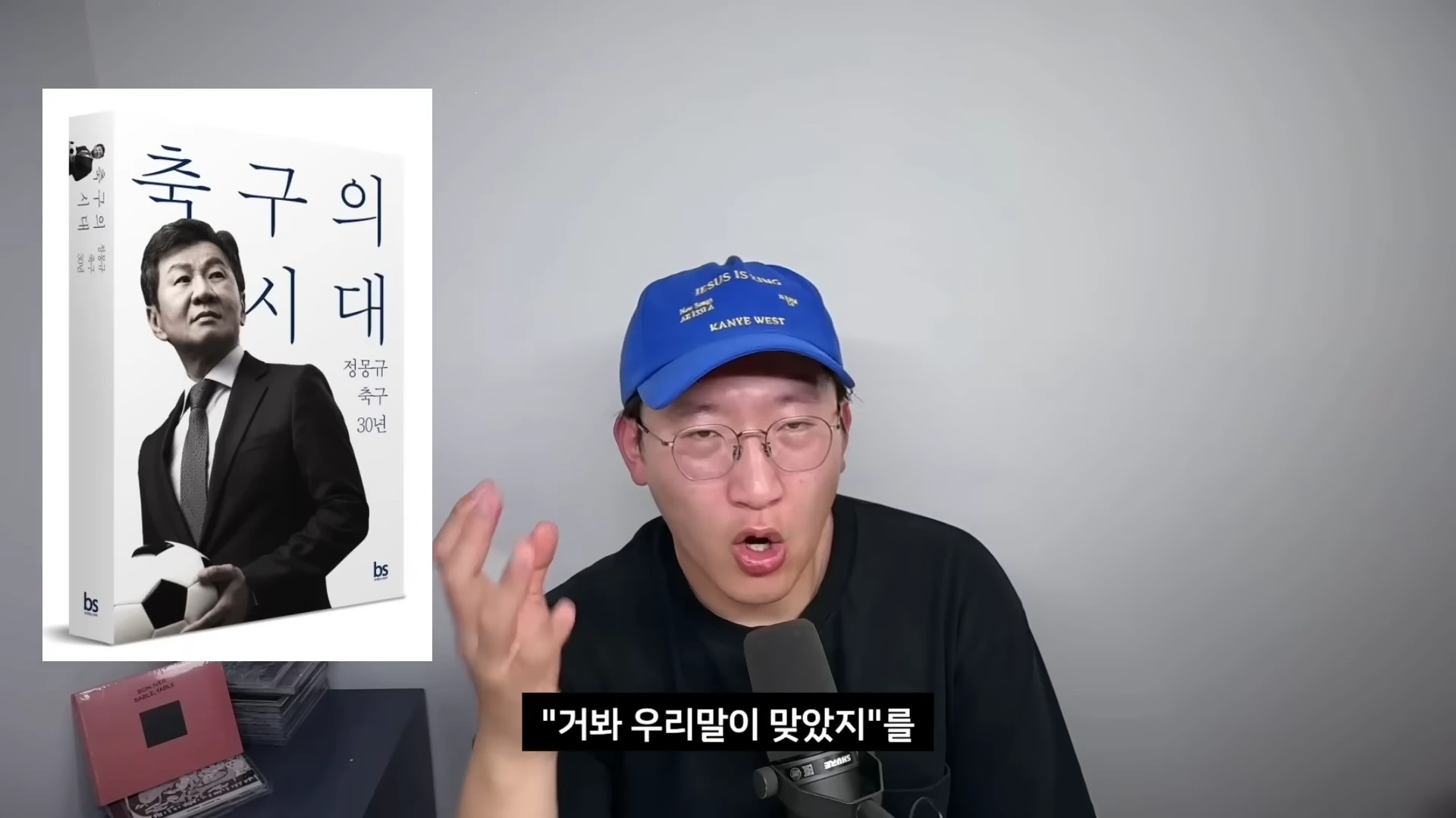 파란색 야구 모자를 쓴 안경 쓴 남성이 마이크 앞에서 이야기하고 있으며, 화면 왼쪽에는 정몽규가 표지에 있는 '축구의 시대'라는 책의 이미지가 배치되어 있습니다.