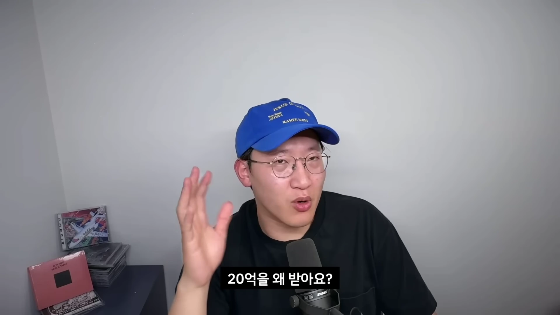 파란색 야구 모자를 쓴 안경을 쓴 남성이 마이크 앞에서 손을 들어 이야기하고 있으며, 화면 하단에는 자막으로 '20억을 왜 받아요?'라는 문구가 적혀 있습니다.
