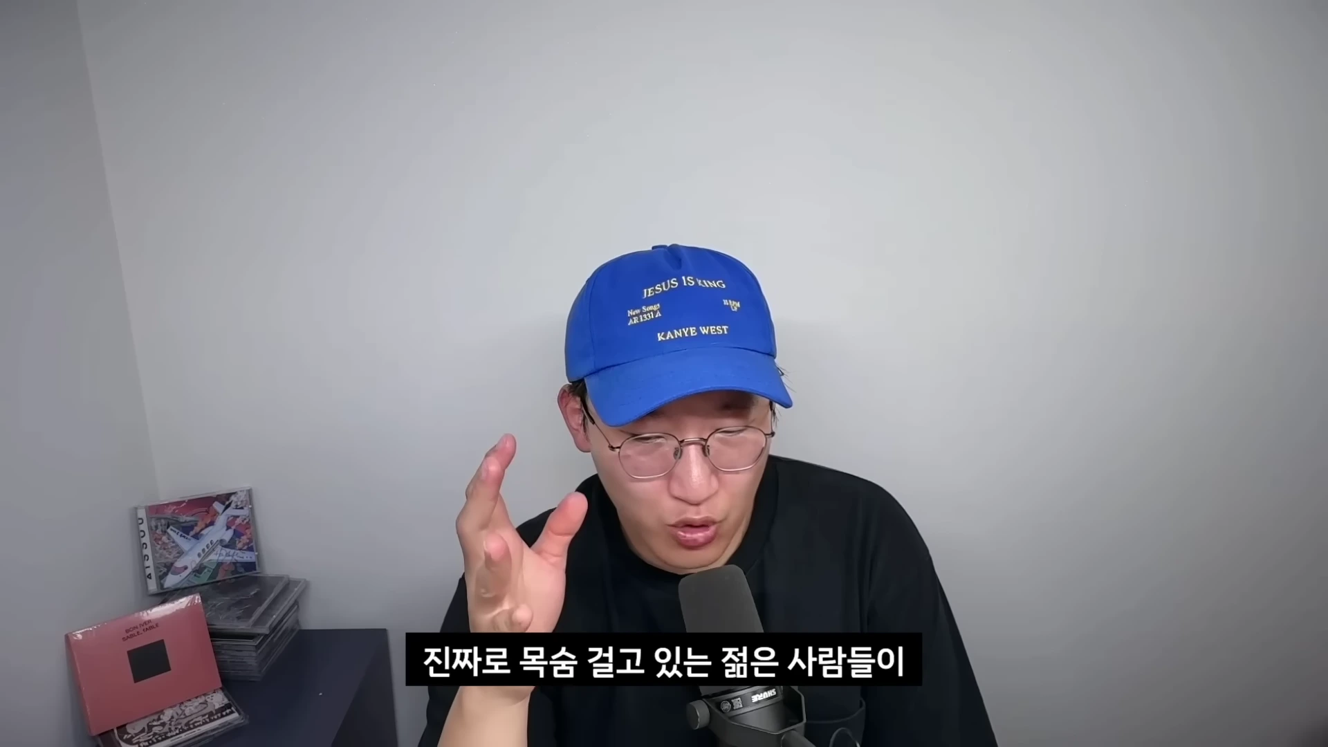 파란색 야구 모자와 안경을 쓴 남성이 마이크 앞에서 손을 들어 대화하고 있는 모습이 담긴 영상 캡처 화면입니다.