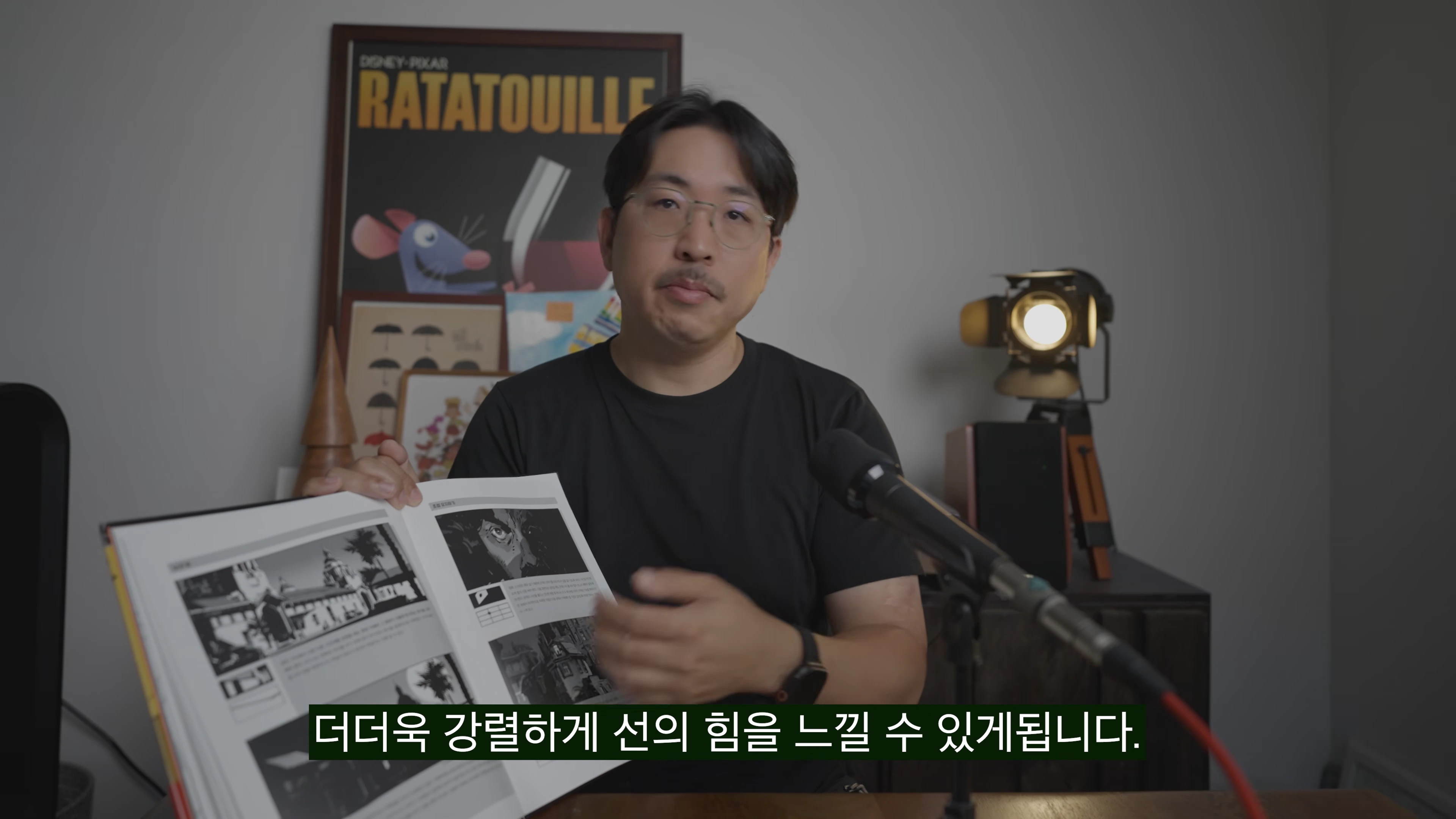 안경을 쓴 한 남성이 검은색 반소매 티셔츠를 입고 책을 펼쳐 보여주고 있으며, 배경에는 영화 라따뚜이 포스터와 조명이 배치된 스튜디오 공간이 보인다.