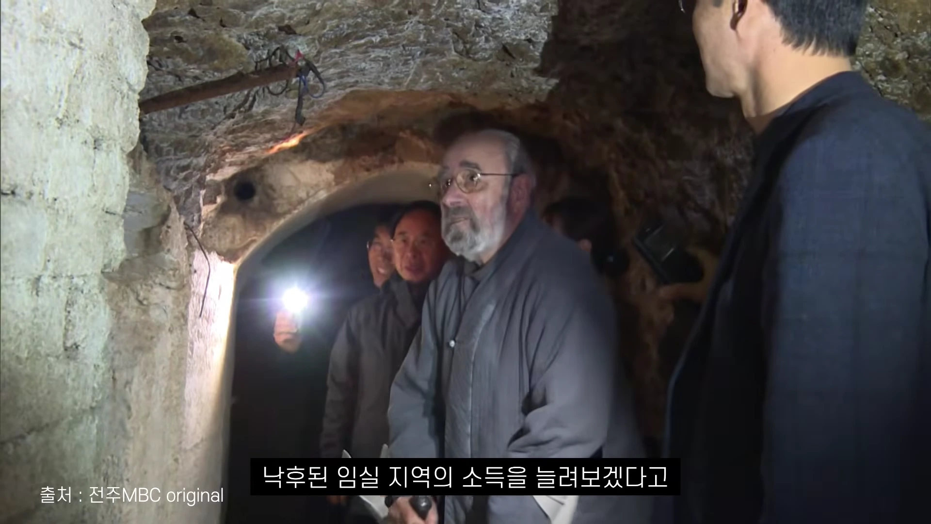 흰 수염을 기른 외국인 신부가 동굴 안에서 사람들과 함께 서 있는 모습