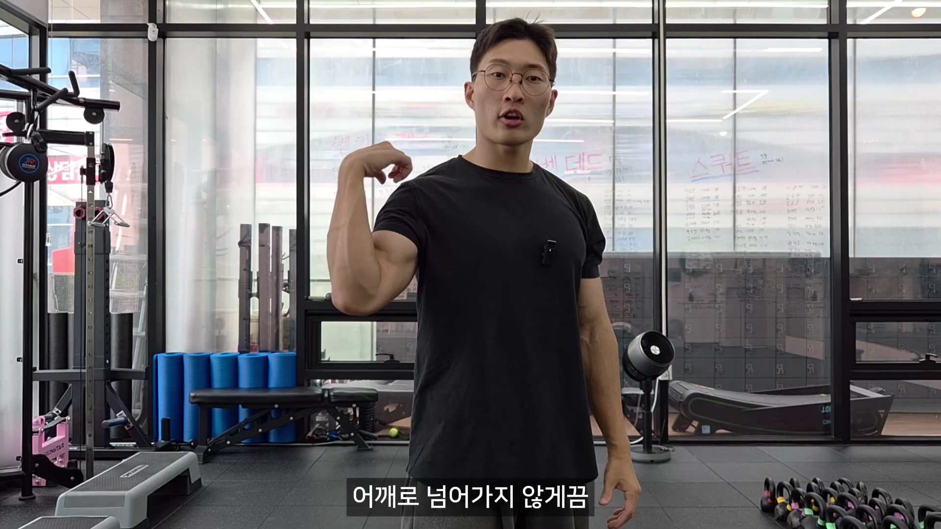 실내 체육관에서 검은색 티셔츠를 입은 남성이 오른손으로 자신의 어깨를 가리키며 설명하고 있다.
