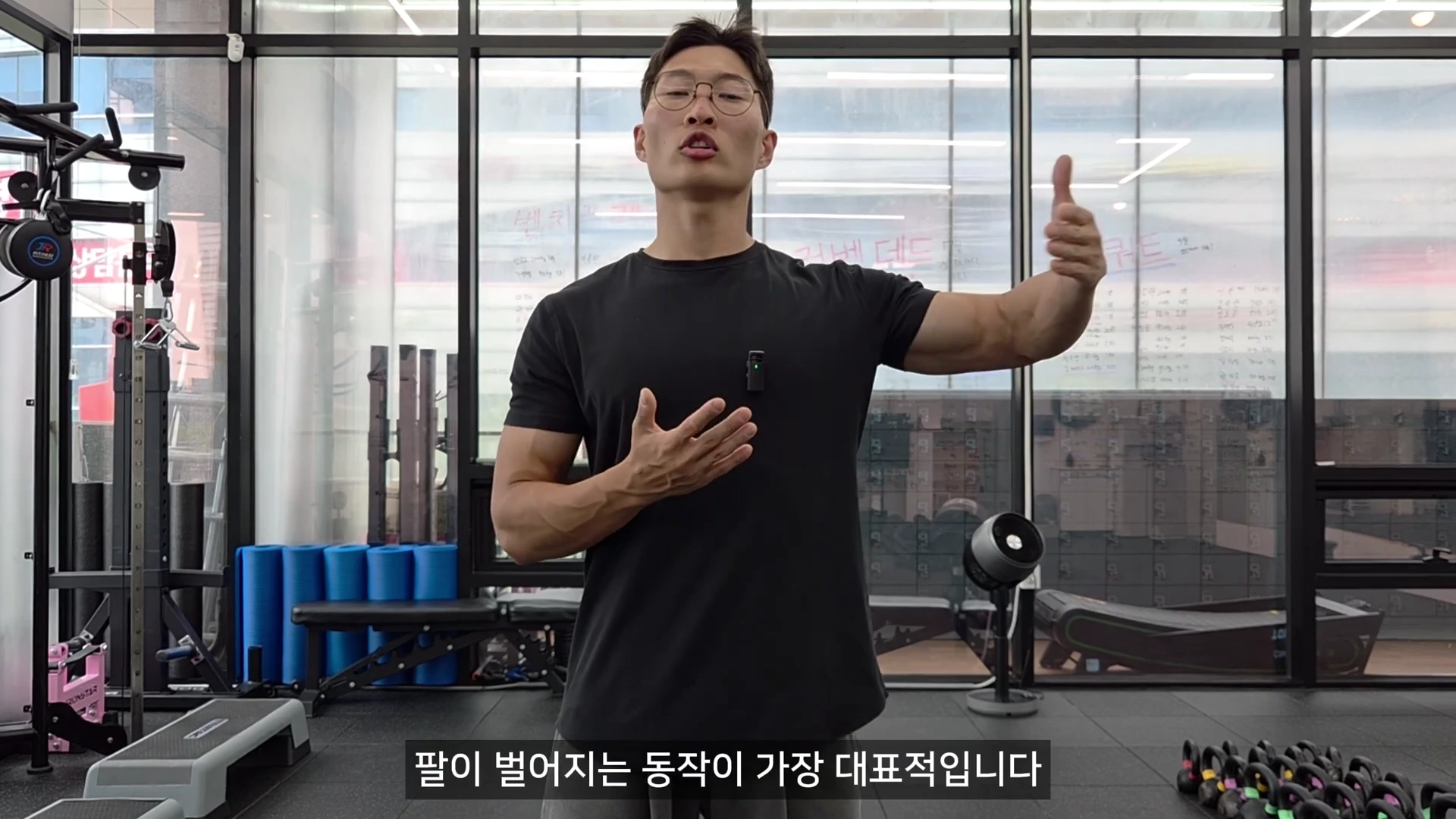 안경을 쓴 남성이 헬스장에서 한쪽 팔을 옆으로 들어 올리는 제스처를 하며 운동 동작을 설명하고 있다.