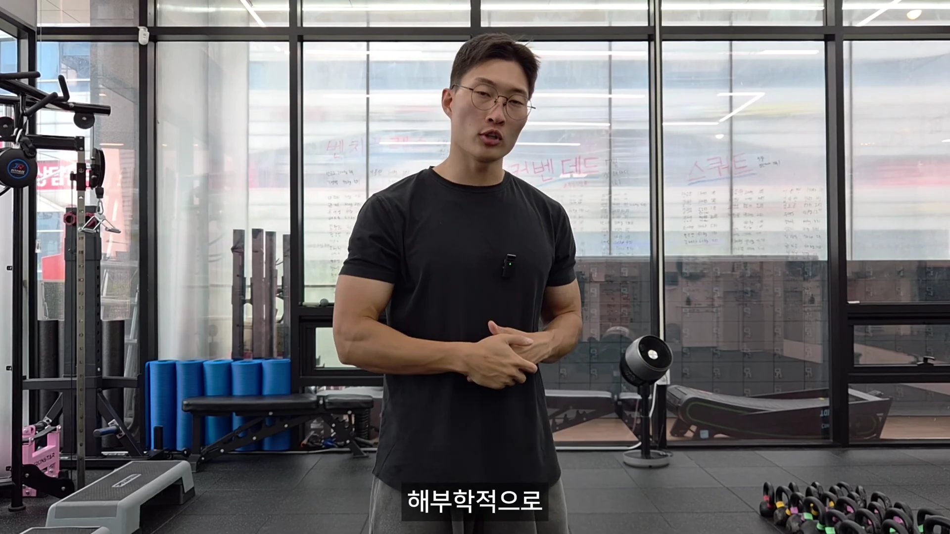 안경을 쓴 남성이 운동복 차림으로 헬스장 내부에서 강의하듯 서서 이야기하고 있습니다.