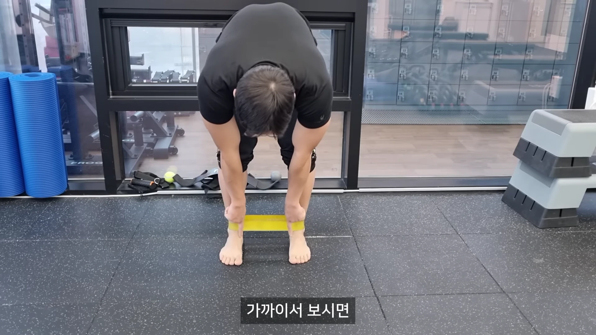 검은색 상의를 입은 남성이 루프 밴드를 발목에 끼우고 상체를 숙인 채 서 있는 모습