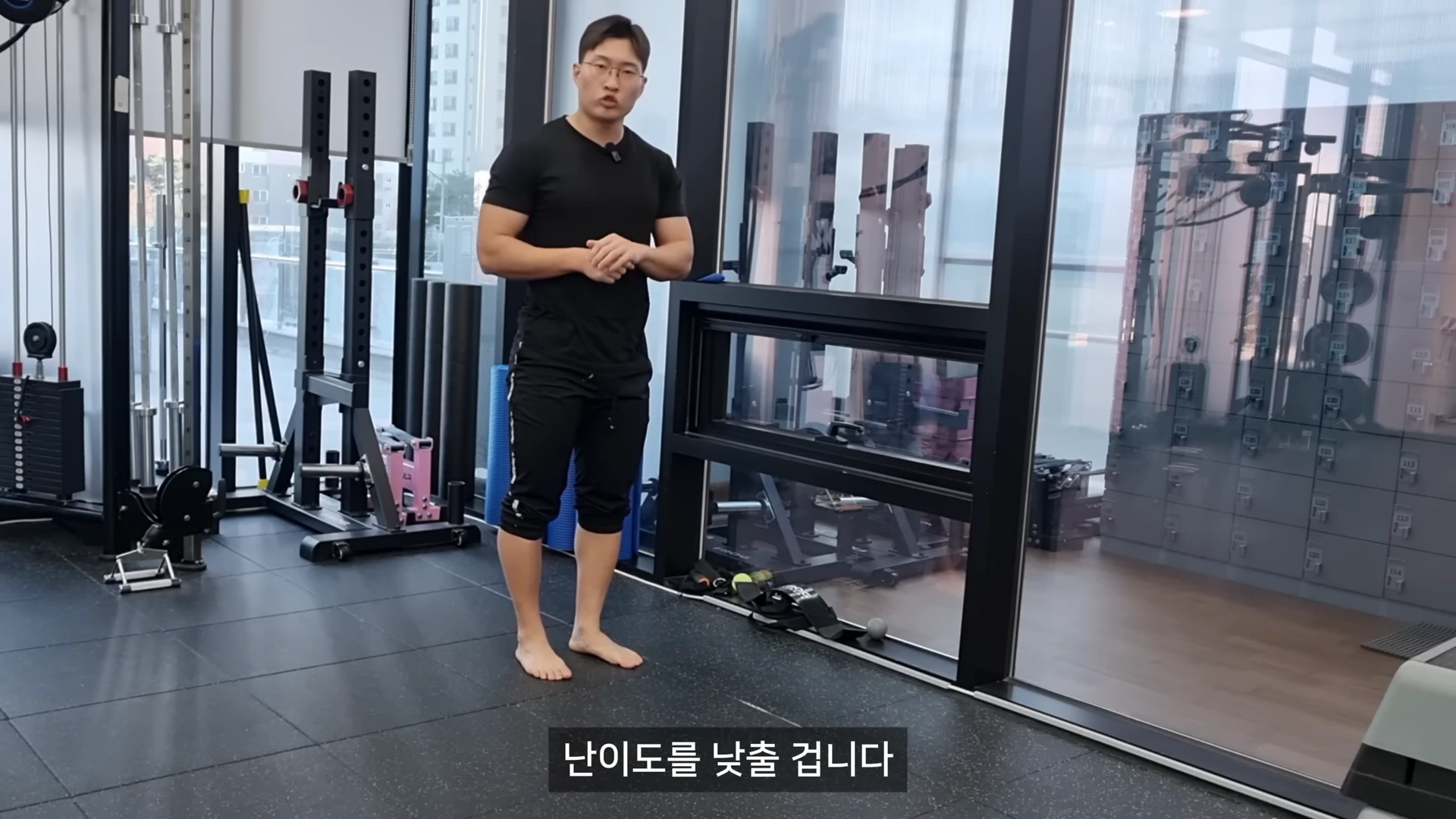 실내 체육관에서 검은색 운동복을 입은 운동 코치가 맨발로 서서 벽을 짚고 원레그 데드리프트 준비 동작을 설명하고 있다.