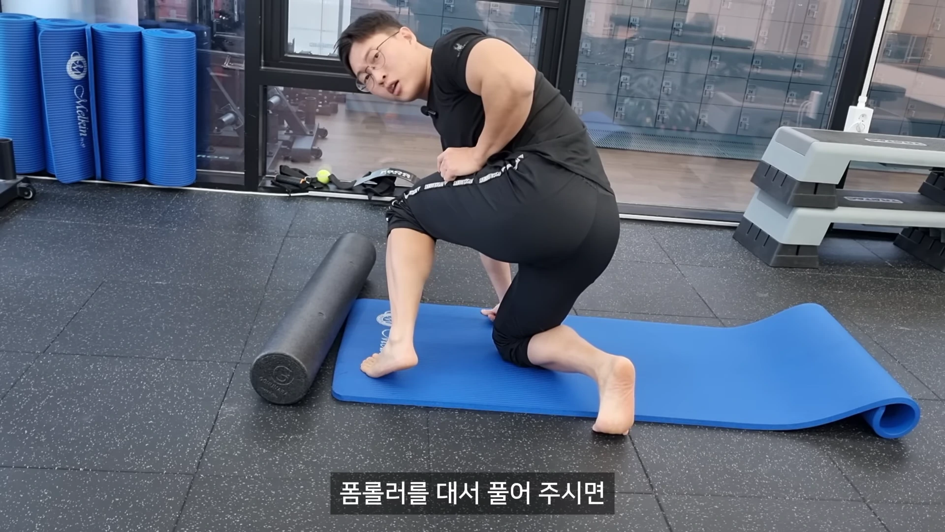 실내 체육관 매트 위에서 한 남성이 폼롤러를 이용해 장딴지 근육을 마사지하는 모습
