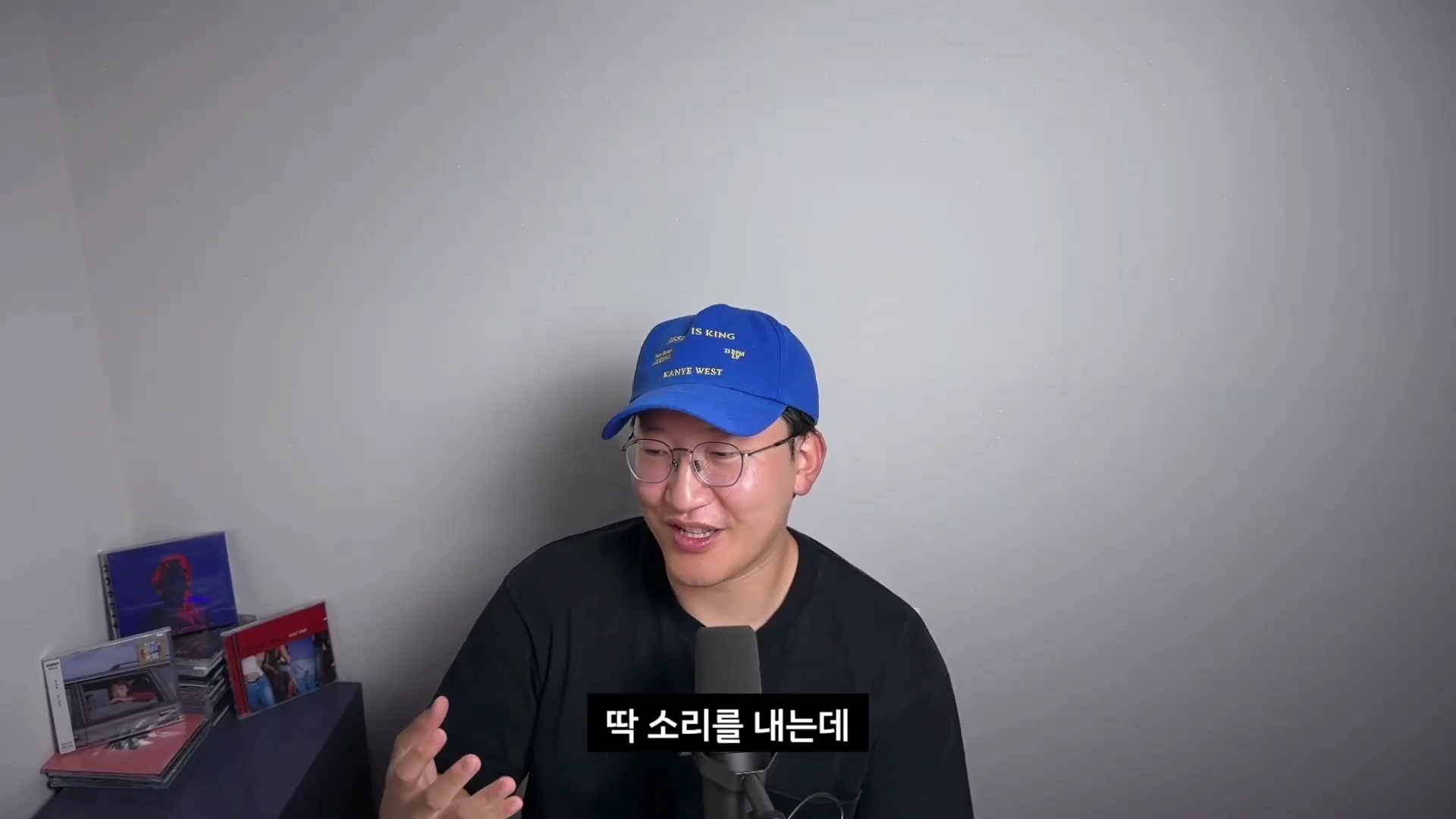 파란색 야구 모자와 안경을 쓴 남성이 마이크 앞에 앉아 손짓하며 이야기를 나누고 있는 모습