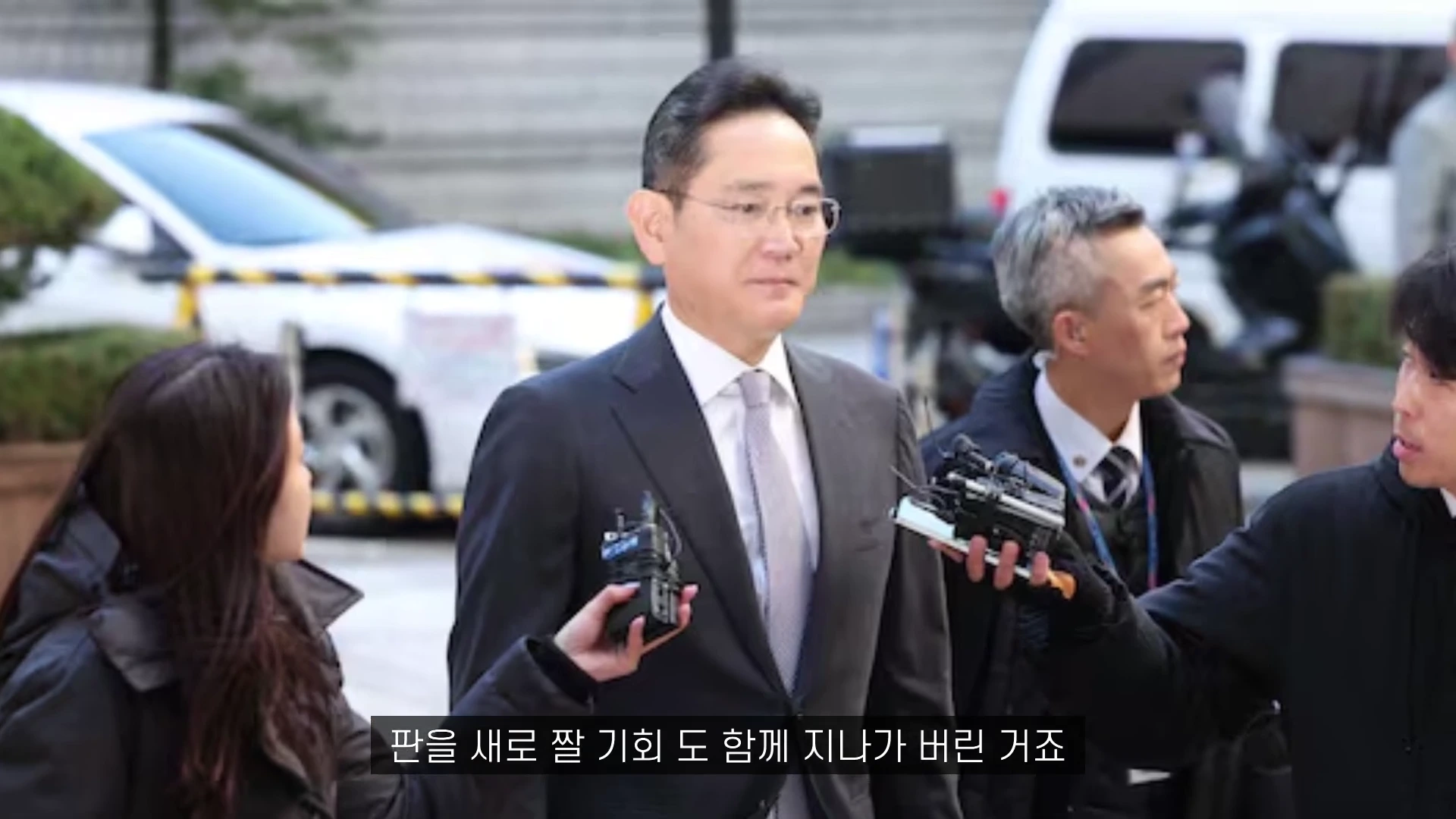 정장을 입은 남성이 여러 기자에게 둘러싸여 마이크를 받고 있는 모습입니다.