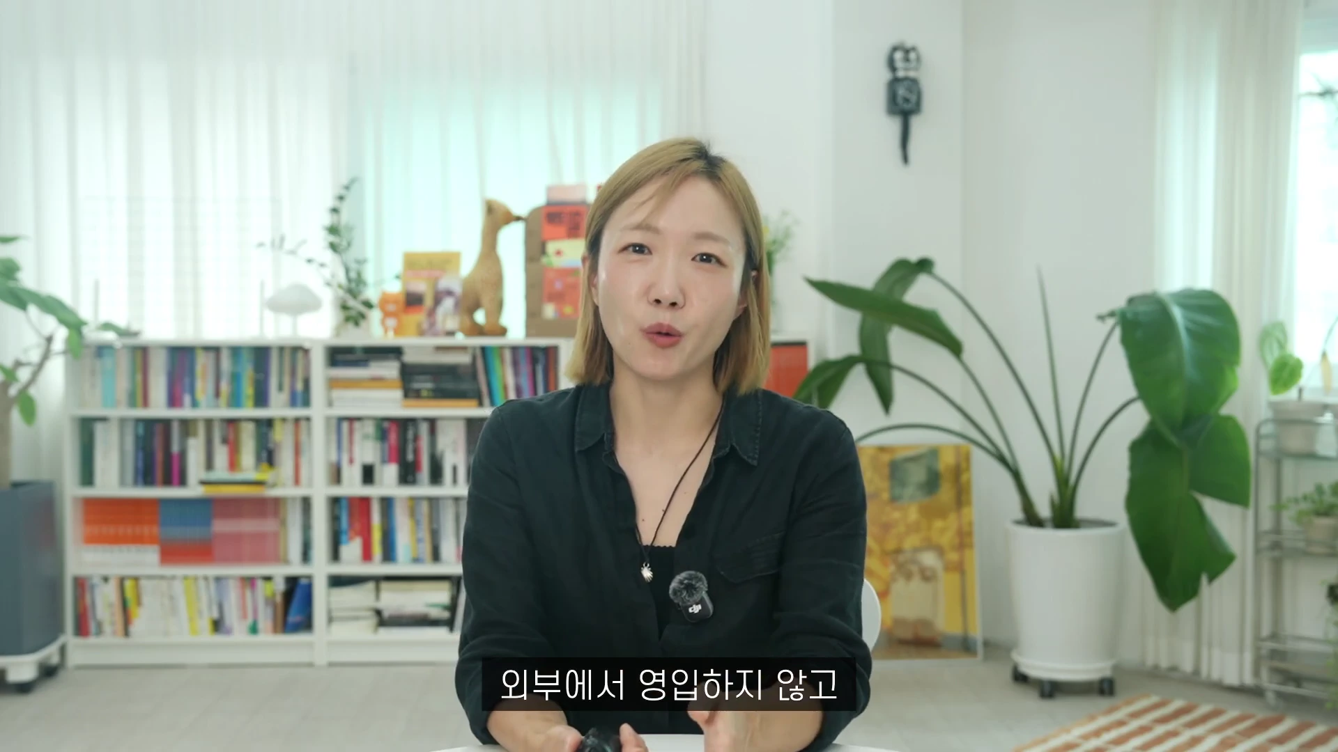 밝은 실내를 배경으로 검은색 셔츠를 입은 단발머리 여성이 카메라를 정면으로 바라보며 앉아 있고 하단에는 자막이 삽입되어 있습니다.