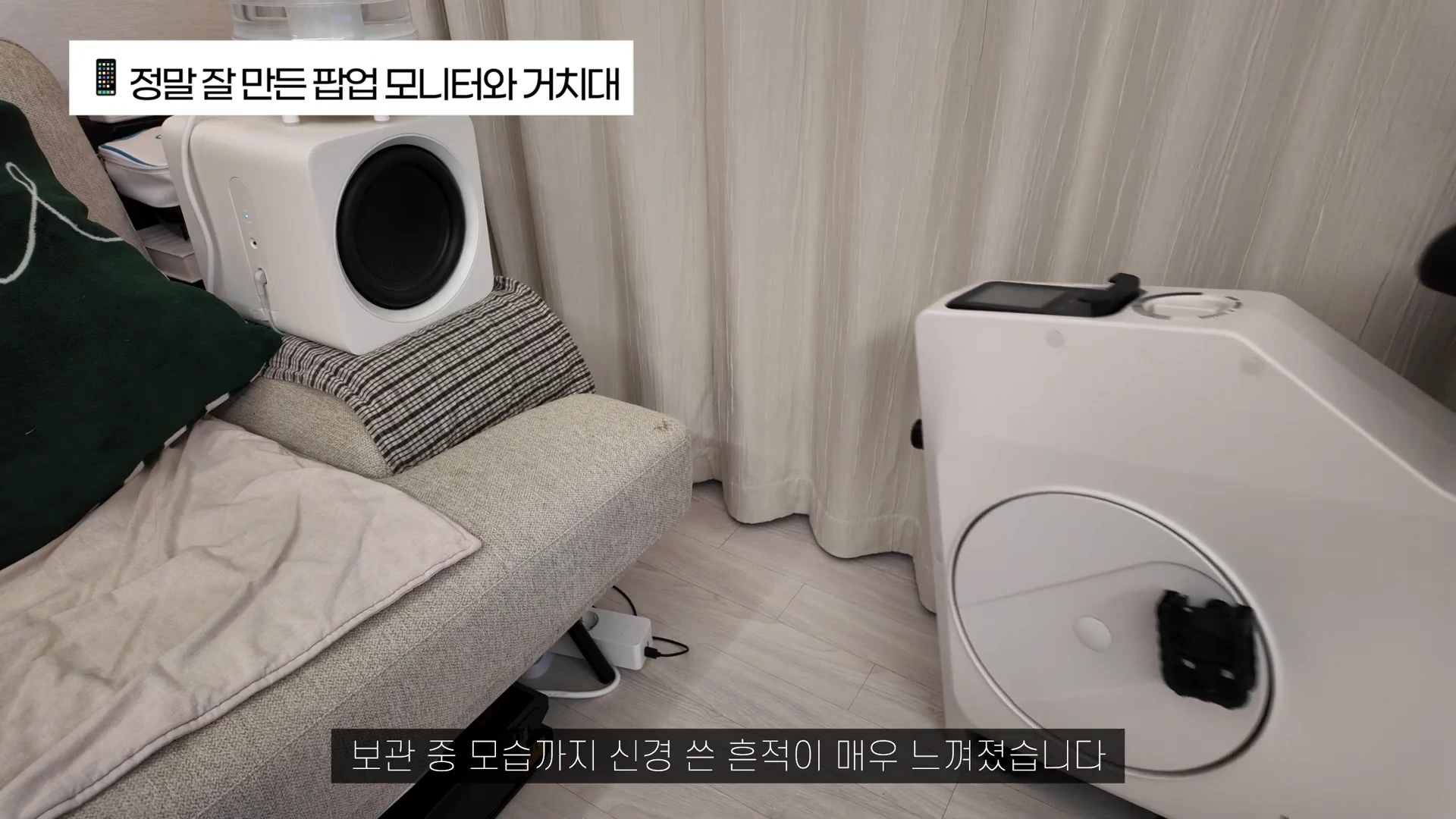 실내에 놓인 흰색 실내자전거와 소파 위의 스피커가 보이며 화면 상단에 '정말 잘 만든 팝업 모니터와 거치대', 하단에 '보관 중 모습까지 신경 쓴 흔적이 매우 느껴졌습니다'라는 자막이 표시된 영상 캡처 화면입니다.