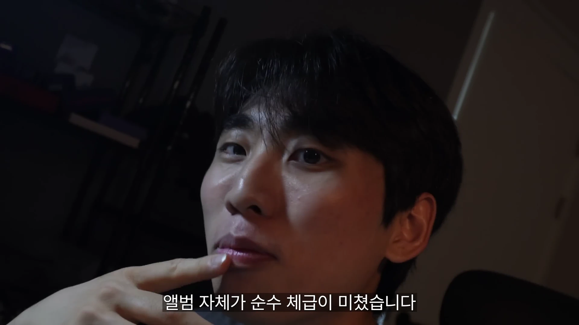 어두운 실내에서 한 남성이 손가락으로 입술을 가리키며 카메라를 응시하고 있으며, 하단에는 한국어 자막이 표시된 영상 화면.