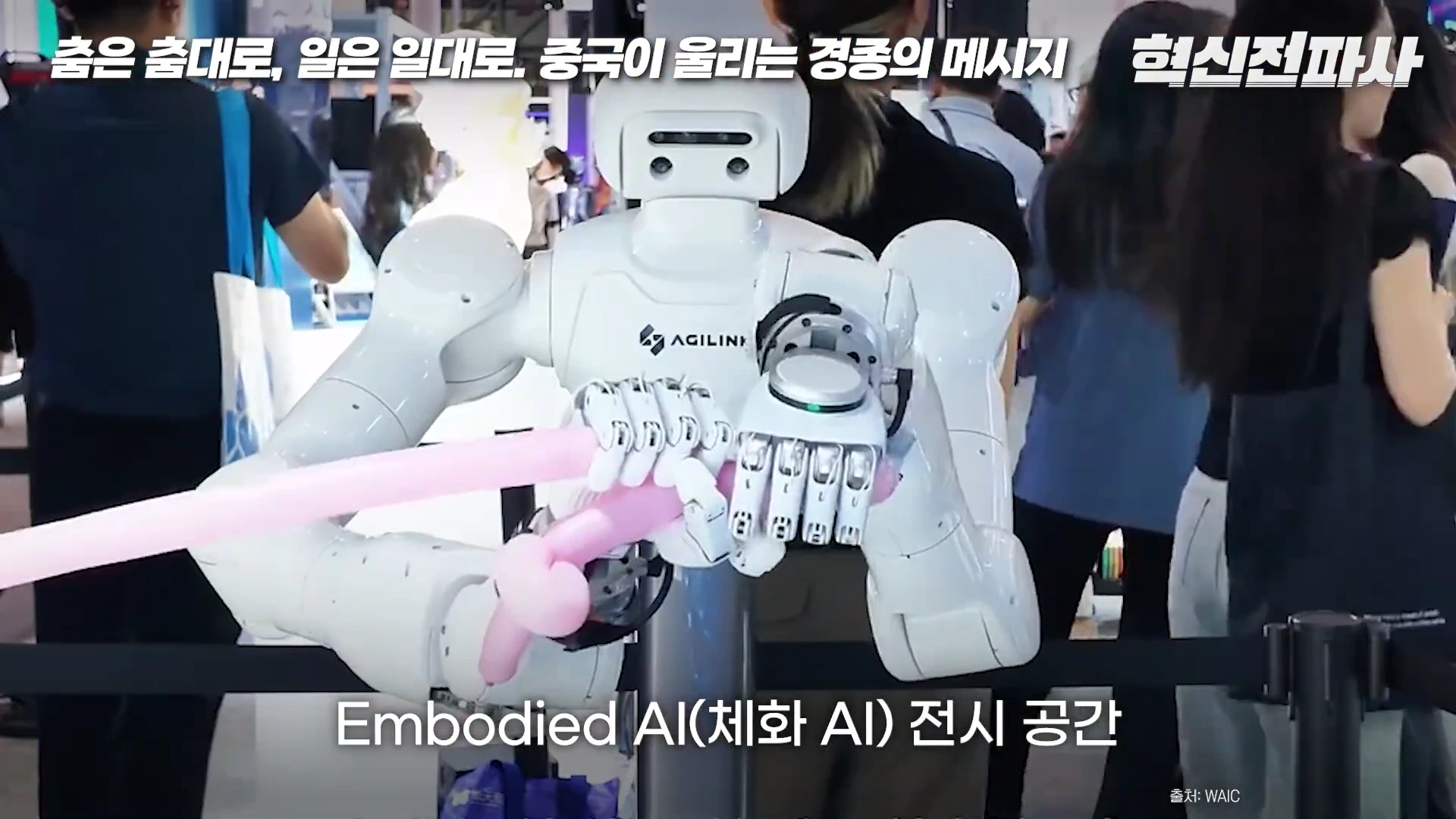 전시장에서 흰색 휴머노이드 로봇이 손으로 분홍색 풍선을 잡고 있는 모습과 배경에 지나가는 관람객들
