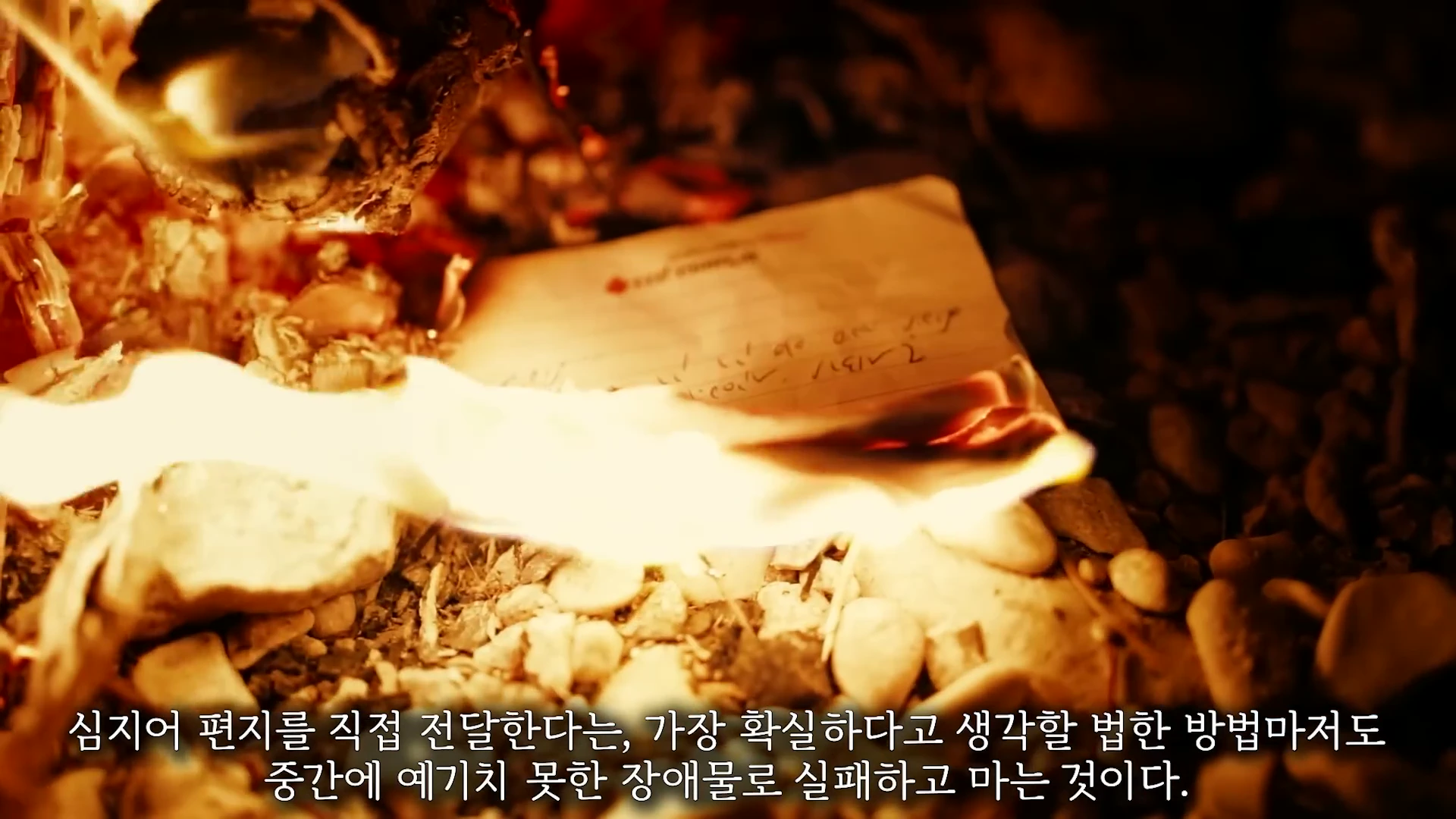 불길에 휩싸여 타들어 가는 종이 편지 한 통이 자갈 바닥 위에 놓여 있는 모습.