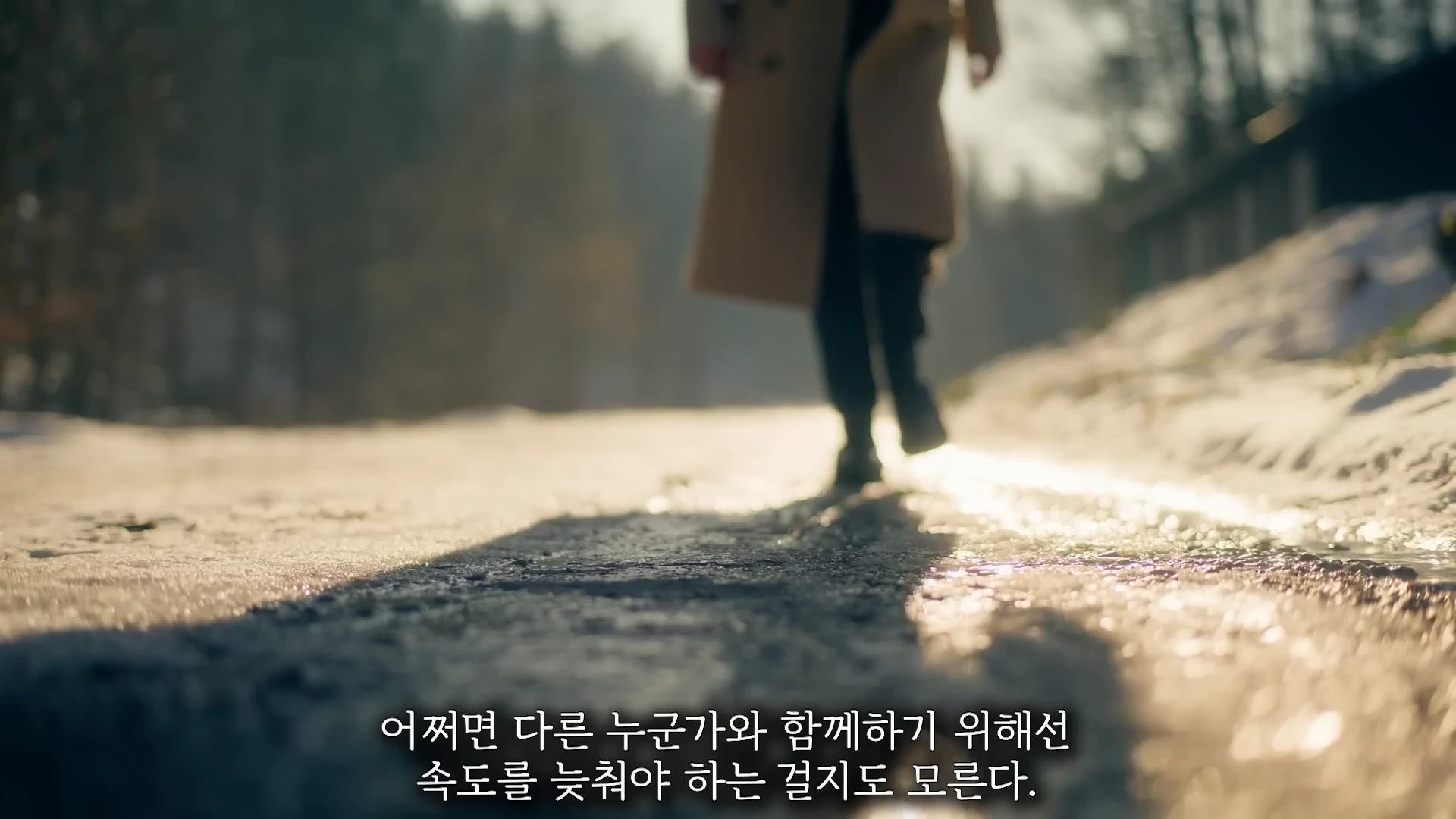 눈이 쌓인 길을 베이지색 코트를 입은 사람이 천천히 걸어가고 있는 뒷모습이 초점이 흐려진 상태로 촬영된 영상 장면입니다. 화면 하단에는 한국어 자막이 적혀 있습니다.