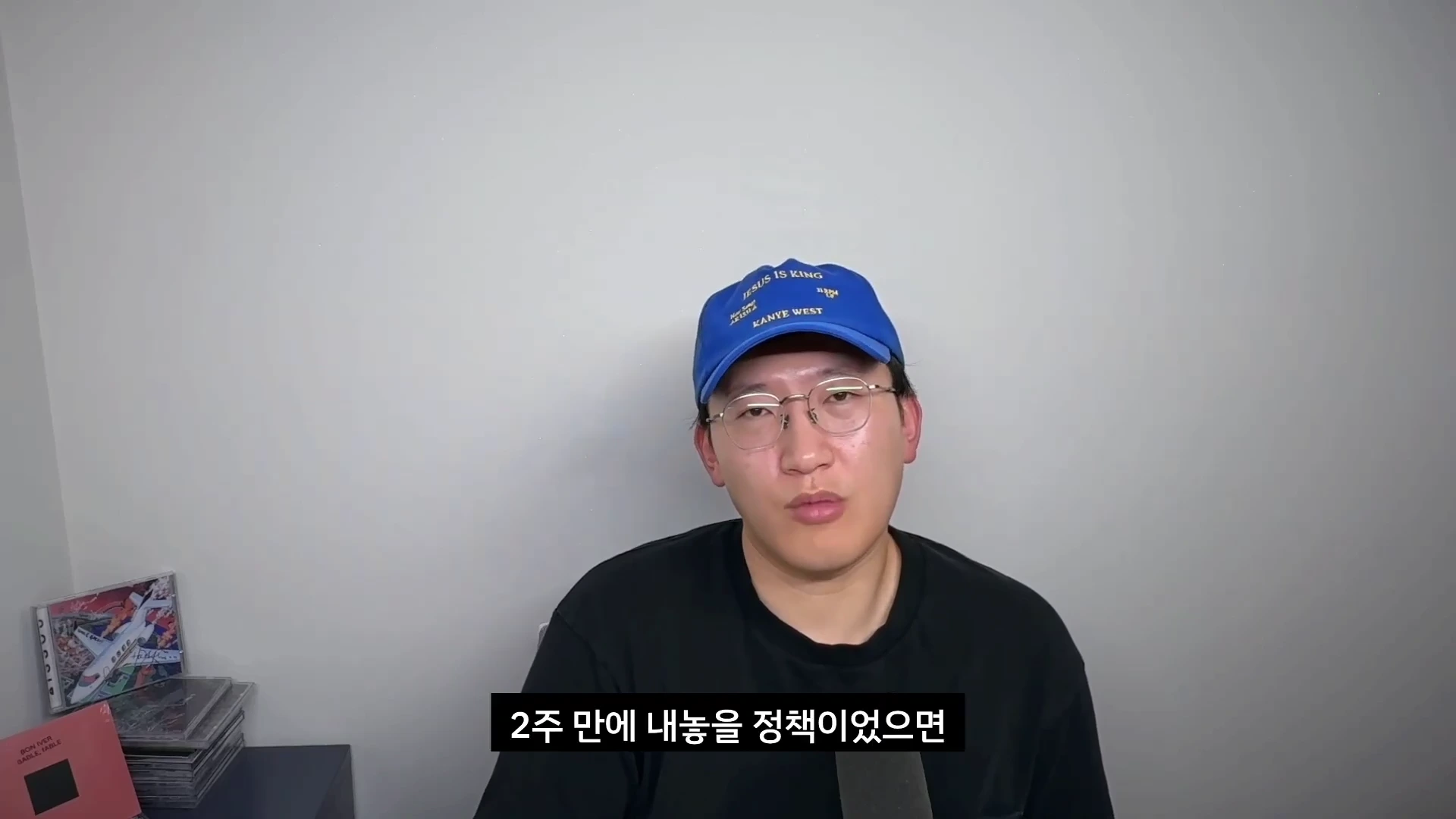 파란색 야구 모자와 안경을 착용한 남성이 흰색 배경 앞에서 카메라를 정면으로 응시하며 이야기하고 있다.