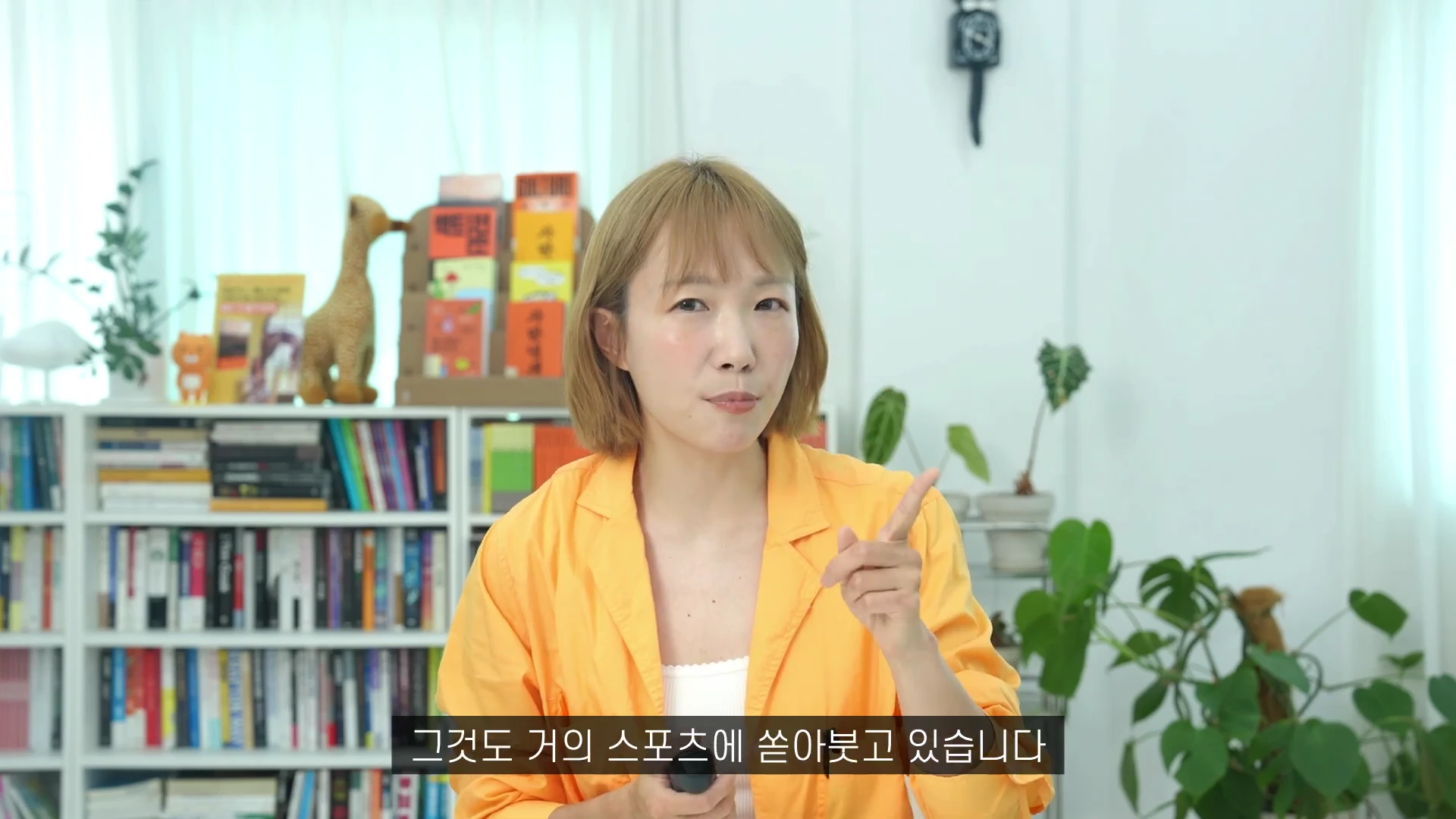 단발머리의 여성이 주황색 셔츠를 입고 밝은 실내 서재를 배경으로 손가락으로 가리키며 이야기하고 있습니다.