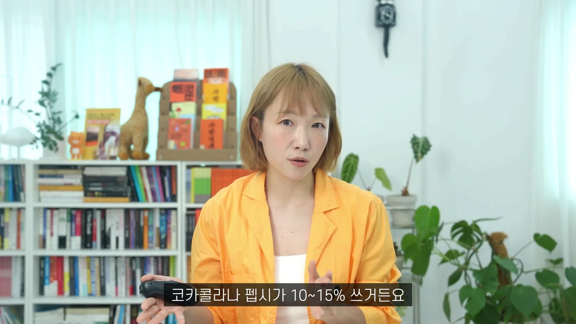 노란색 재킷을 입은 단발머리 여성 진행자가 책장이 있는 밝은 실내에서 무언가를 설명하고 있으며, 화면 하단에는 코카콜라나 펩시의 마케팅 비용에 관한 자막이 표시되어 있습니다.