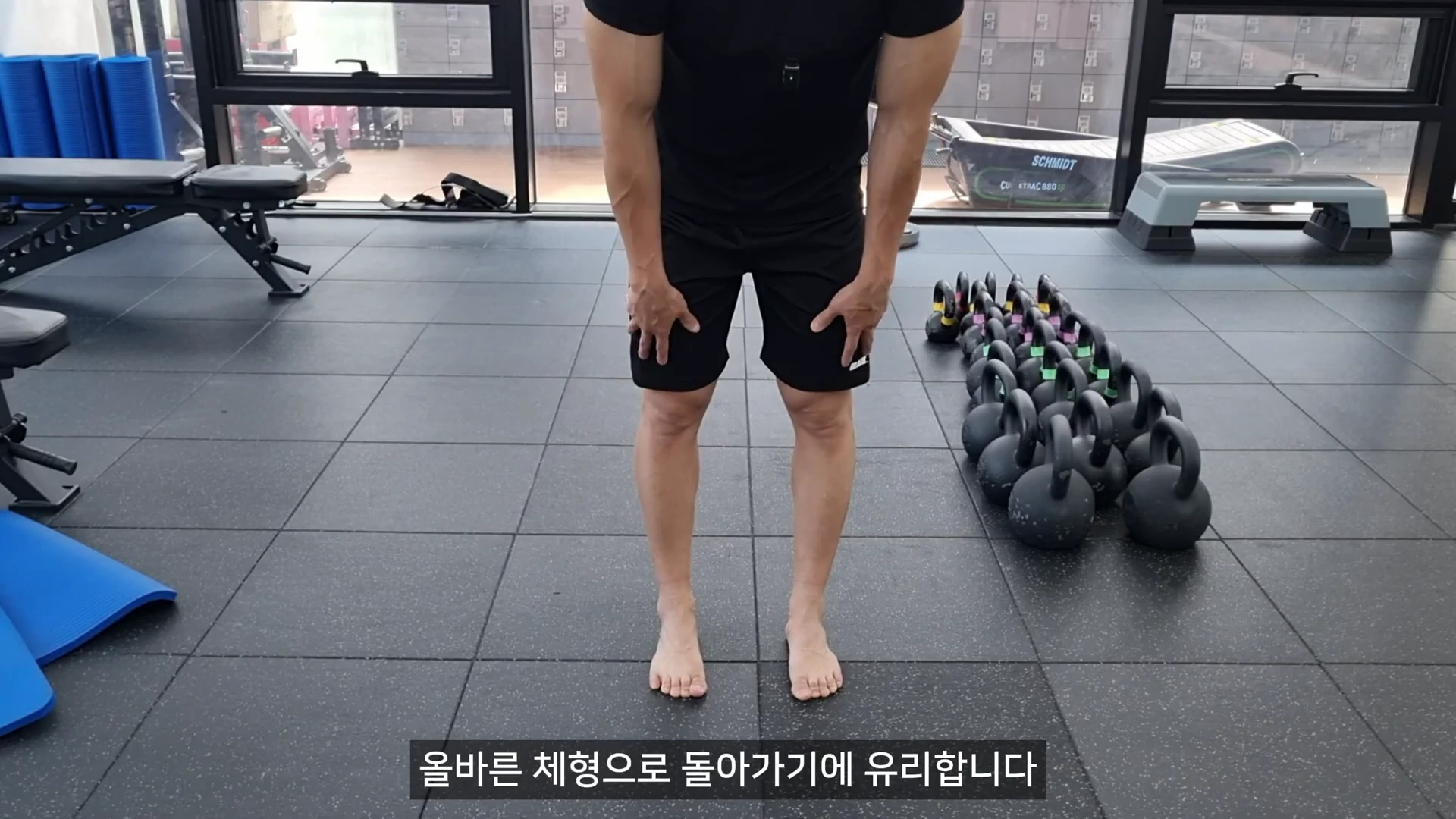 피트니스 센터 바닥에 맨발로 서 있는 사람의 하체 모습으로, 다리를 11자로 나란히 정렬하고 무릎 위에 양손을 가볍게 얹고 있는 전신 운동 영상의 한 장면입니다.