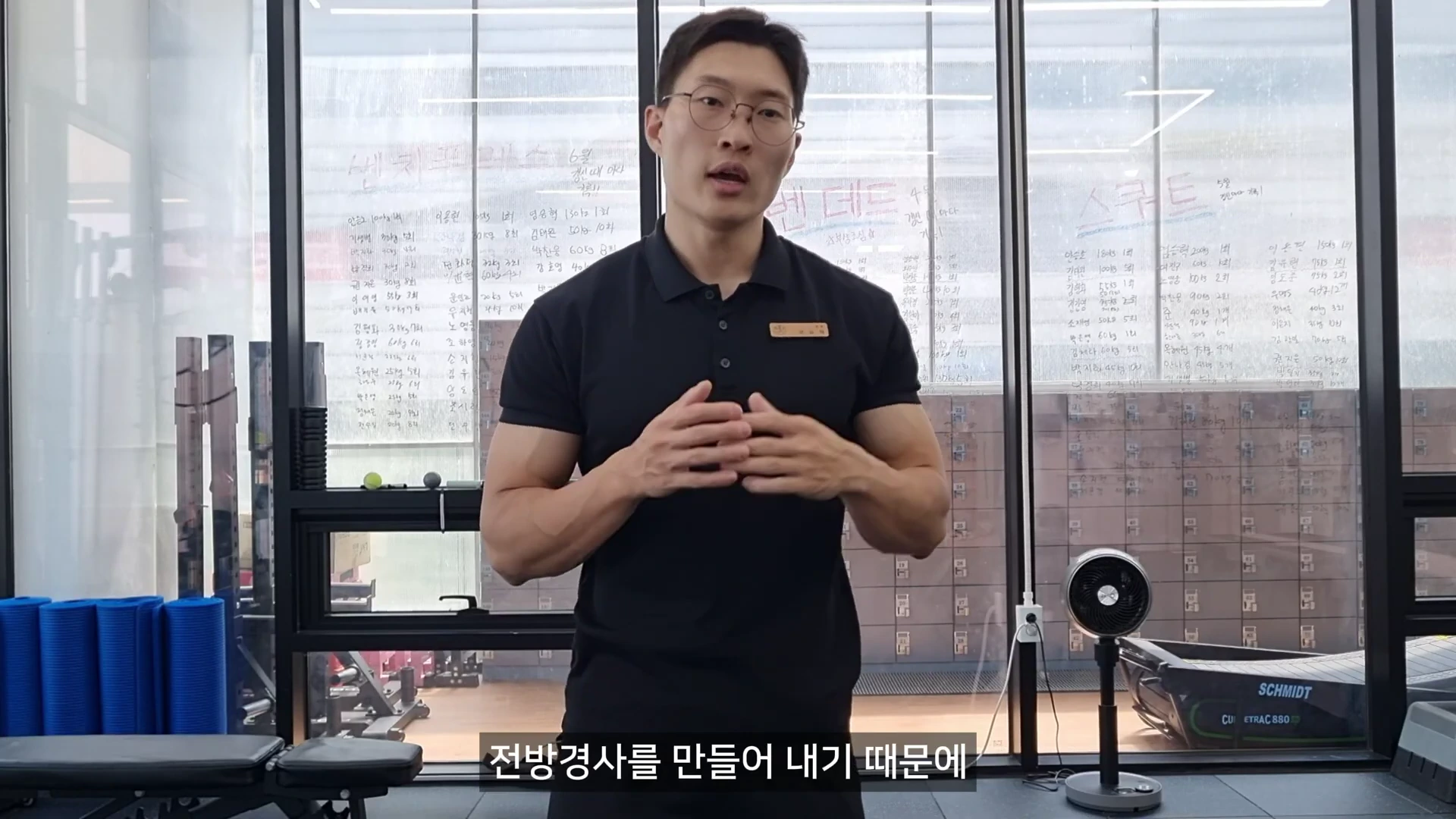 안경을 쓴 남성이 운동 센터에서 상체를 세우고 서서 설명하고 있는 모습이며, 뒷배경의 유리창에는 각종 기록이 적혀 있습니다.