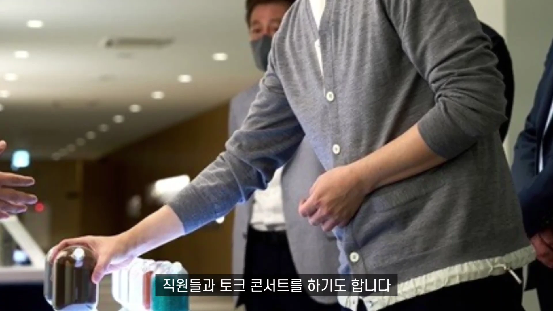 회색 카디건을 입은 남성이 사무 공간에서 전시된 샘플을 살피고 있으며 그 아래로 자막이 배치되어 있다.