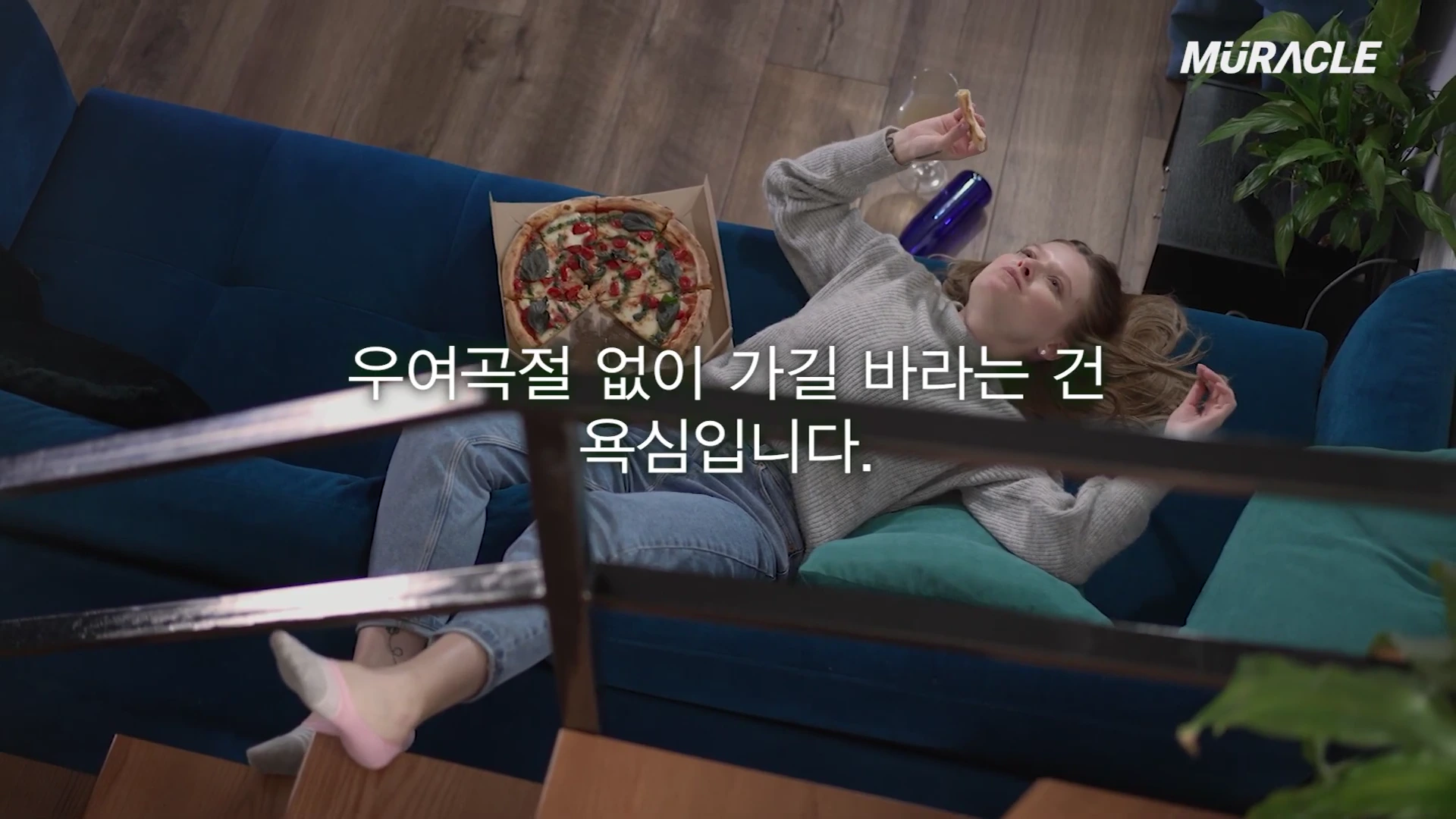 푸른색 소파 위에 한 여성이 누워 피자를 먹으며 쉬고 있는 모습이 위에서 내려다보는 구도로 촬영되었으며, 화면 중앙에는 한국어 자막이 입혀져 있습니다.
