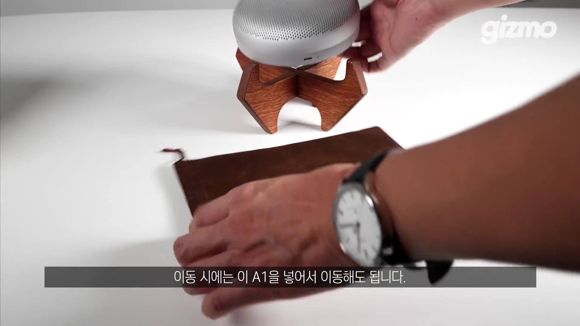 나무 소재의 X자형 스피커 거치대 위에 은색 원형 스피커가 놓여 있고, 손이 그 옆에 갈색 파우치를 두고 있는 모습