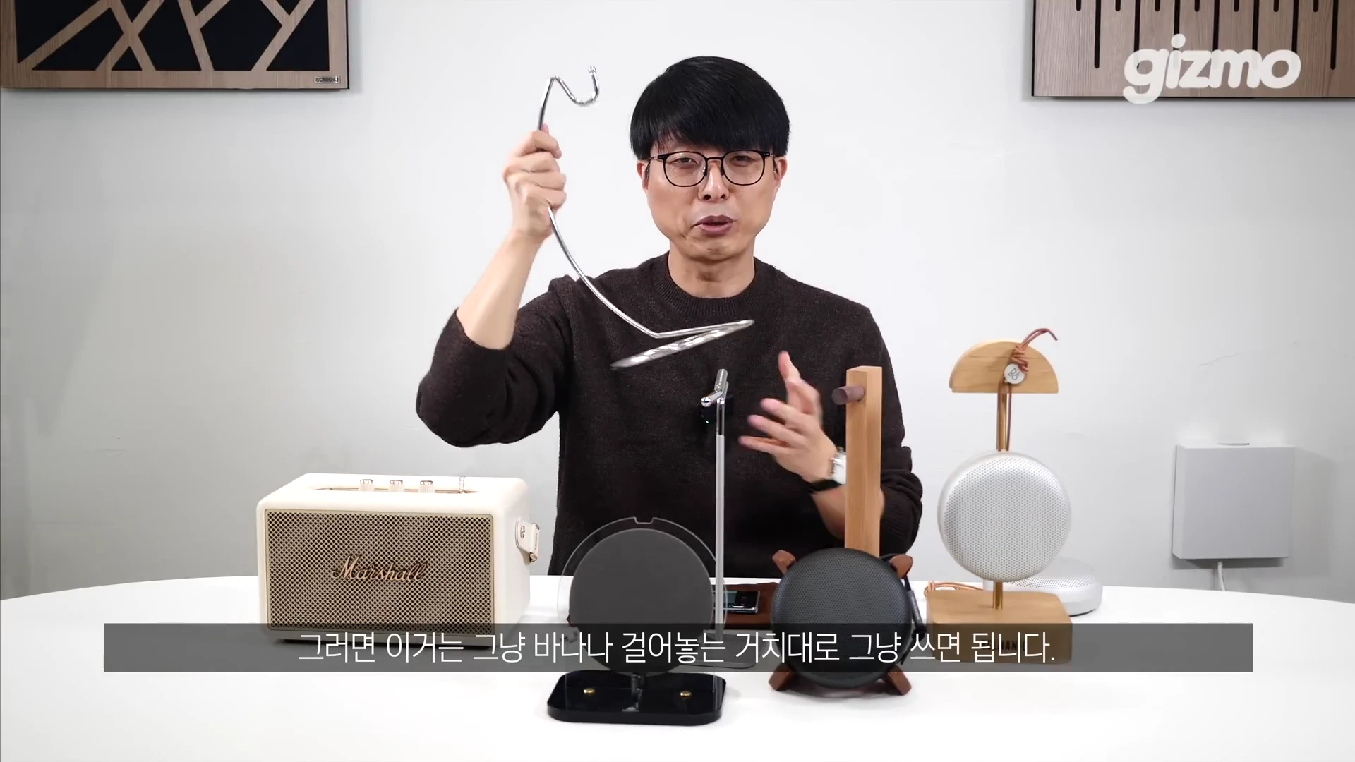 안경을 쓴 남성이 스튜디오 테이블 앞에 앉아 한 손에는 금속제 바나나 걸이를 들어 보이고 있으며, 테이블 위에는 스피커와 다양한 형태의 스탠딩 거치대가 놓여 있다.