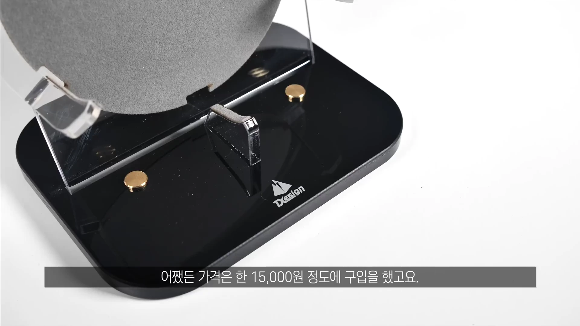투명한 아크릴 소재의 거치대가 검은색 받침대 위에 고정되어 있고, 그 위에 금색 나사 장식과 영어 브랜드 로고가 새겨진 모습이 담긴 클로즈업 사진입니다.