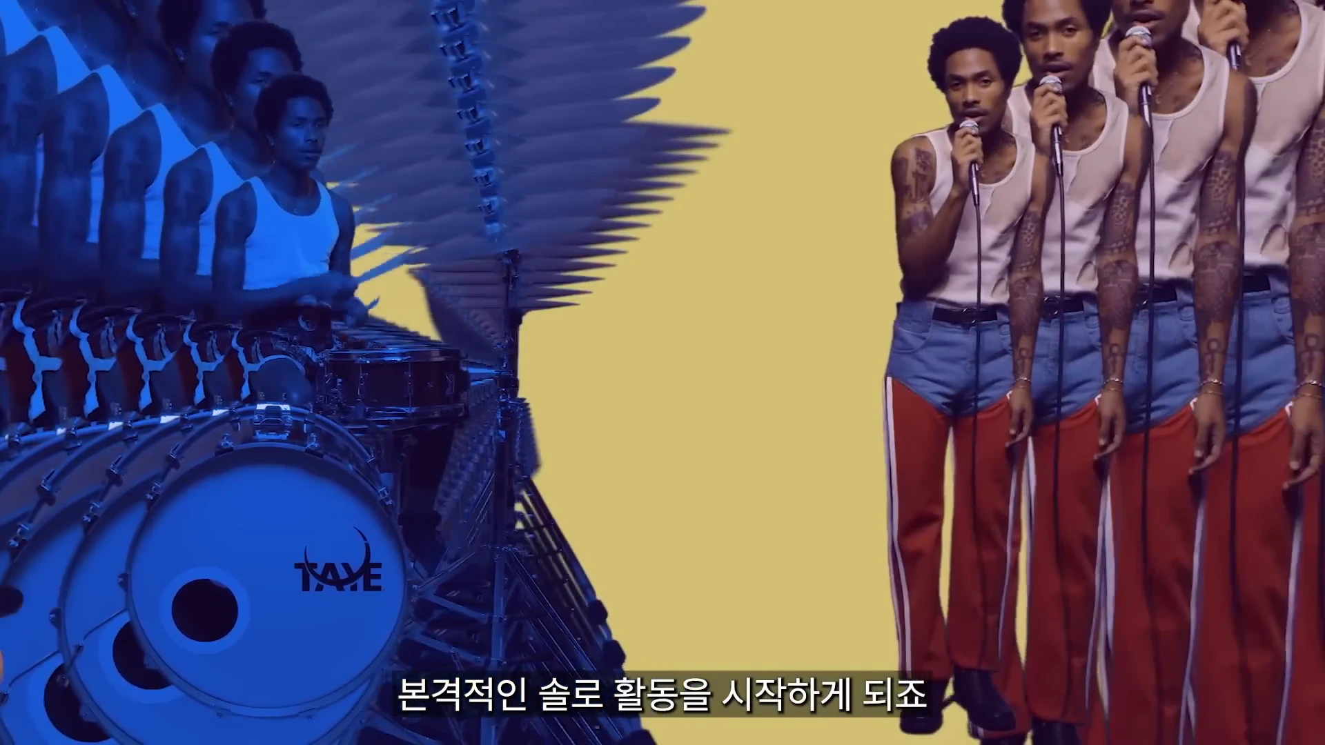 흰색 민소매 셔츠와 독특한 바지를 입은 한 남성이 여러 번 복제되어 드럼을 연주하고 마이크를 잡고 노래하는 모습이 담긴 화면