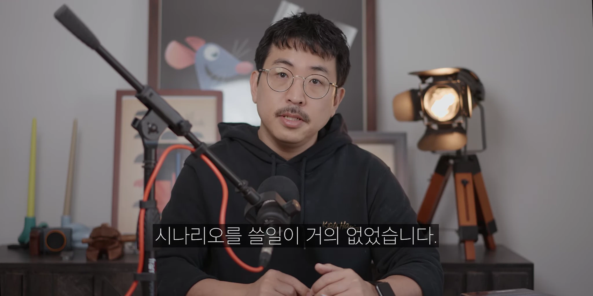 안경을 쓴 남성이 마이크 앞에 앉아 이야기하고 있으며 뒤편에는 캐릭터 액자와 조명이 놓여 있다.