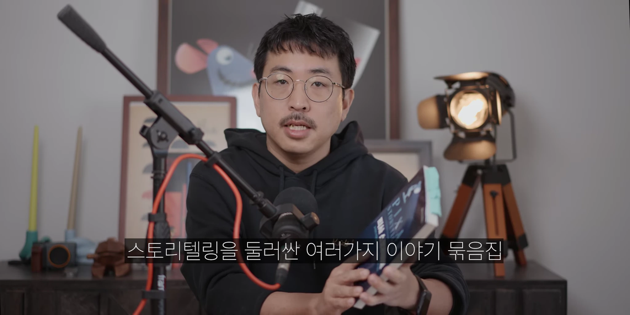 안경을 쓴 한 남성이 검은색 후드티를 입고 손에 책을 든 채 마이크 앞에서 이야기하고 있으며, 배경에는 액자와 조명이 보입니다.