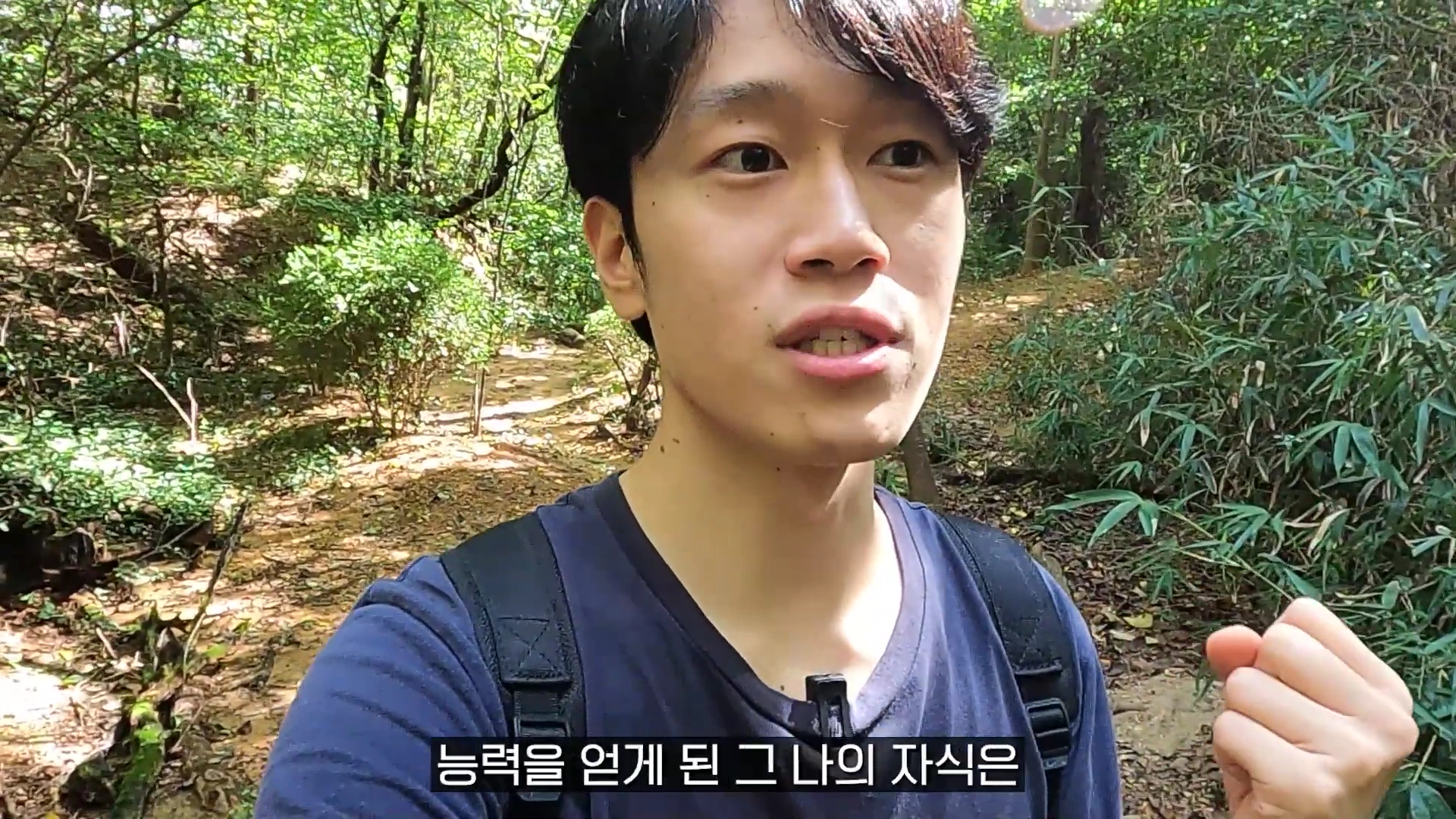 숲길을 배경으로 검은색 티셔츠를 입은 젊은 남성이 카메라를 보며 손짓을 섞어 이야기하고 있다.