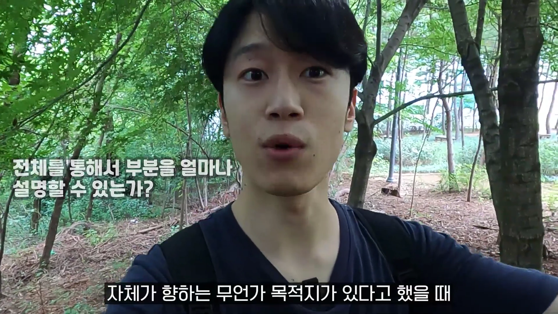 나무가 우거진 숲길을 배경으로 한 남성이 화면을 응시하며 이야기하고 있으며, 화면 위에는 '전체를 통해서 부분을 얼마나 설명할 수 있는가?'와 '자체가 향하는 무언가 목적지가 있다고 했을 때'라는 한국어 자막이 입혀져 있습니다.