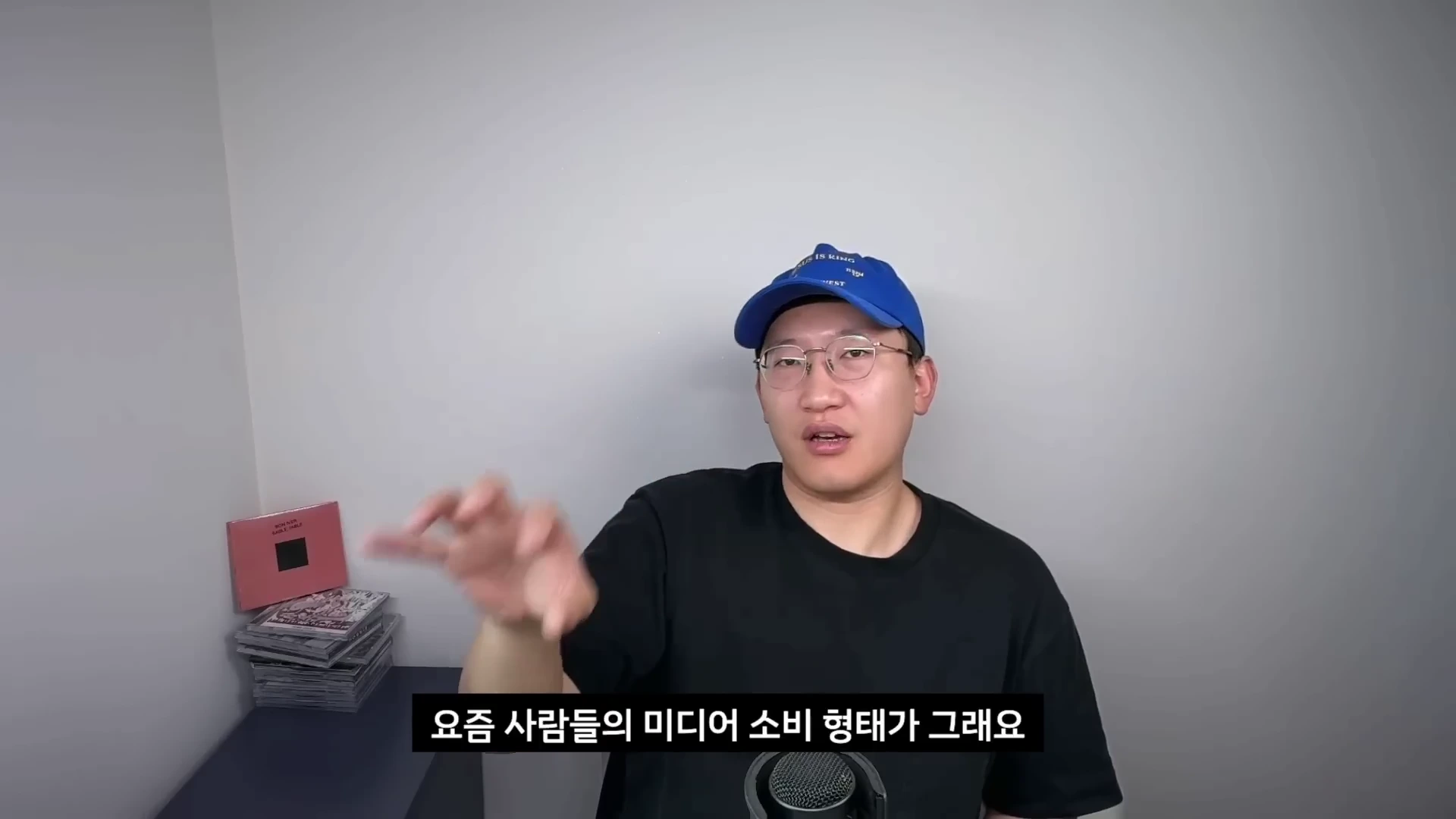 파란색 야구 모자와 안경을 착용한 남성이 마이크 앞에서 손동작을 하며 이야기하고 있고, 하단에는 '요즘 사람들의 미디어 소비 형태가 그래요'라는 자막이 표시되어 있습니다.