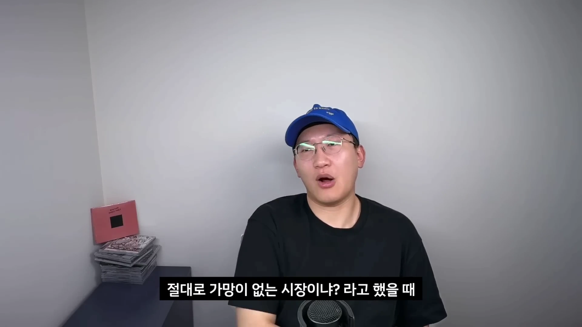 파란색 야구 모자와 안경을 착용한 남성이 마이크 앞에 앉아 이야기를 하고 있는 모습.