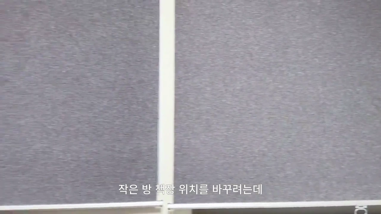 두 개의 회색 패널 벽면과 그 위에 '작은 방 책장 위치를 바꾸려는데'라는 한국어 자막이 겹쳐져 있는 영상 화면.