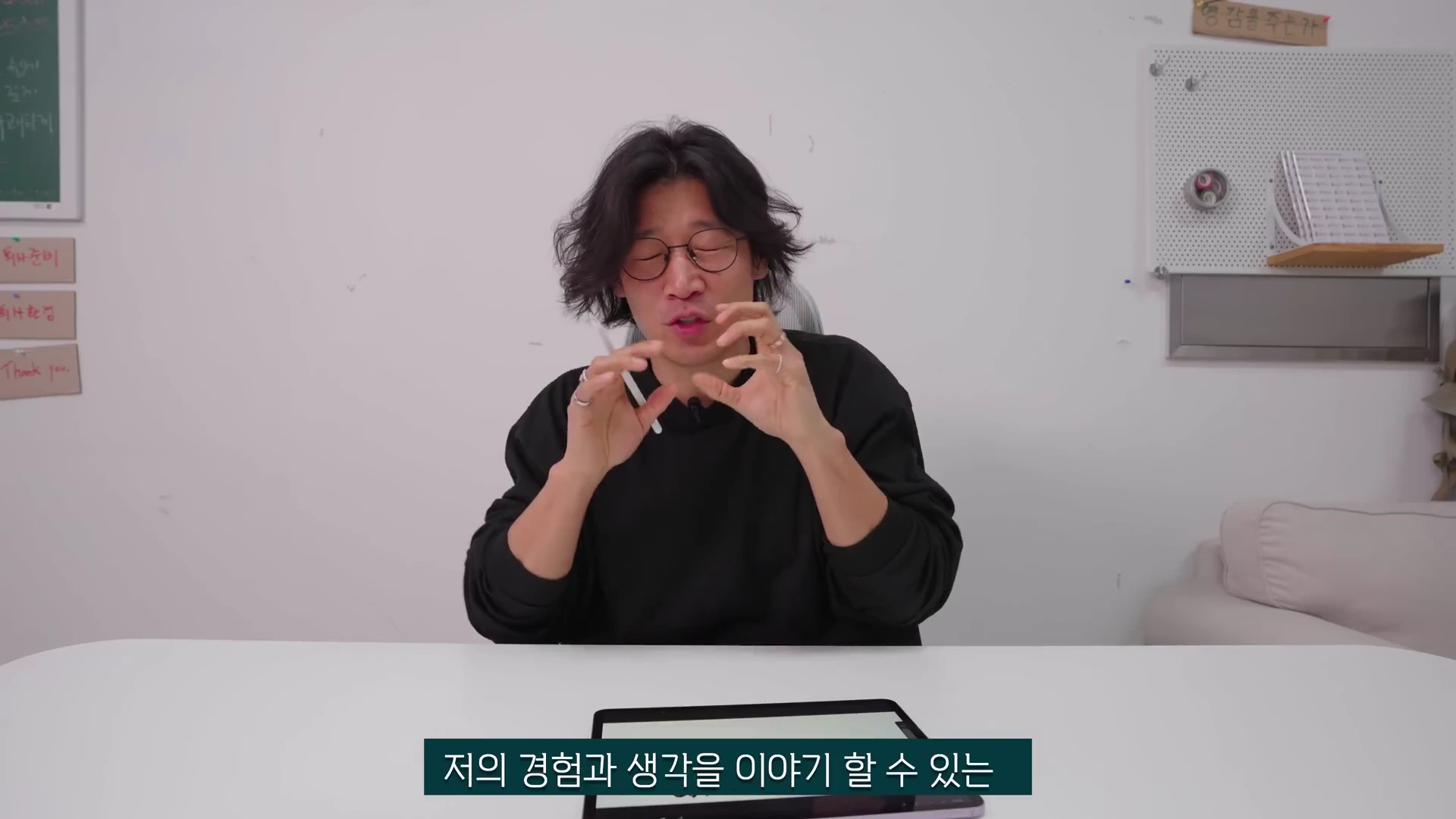흰 배경의 방 안에서 검은색 긴팔 티셔츠를 입고 안경을 쓴 남성이 테이블 앞에 앉아 손동작을 하며 말을 하고 있는 모습