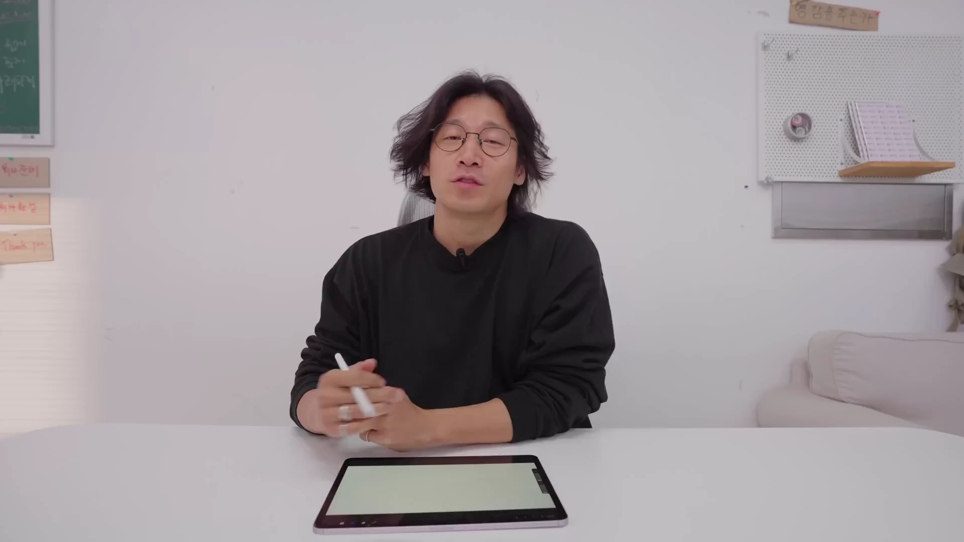 흰색 배경의 밝은 스튜디오에서 검은색 긴팔 티셔츠를 입고 안경을 쓴 중년 남성이 태블릿을 앞에 두고 앉아 이야기하는 모습