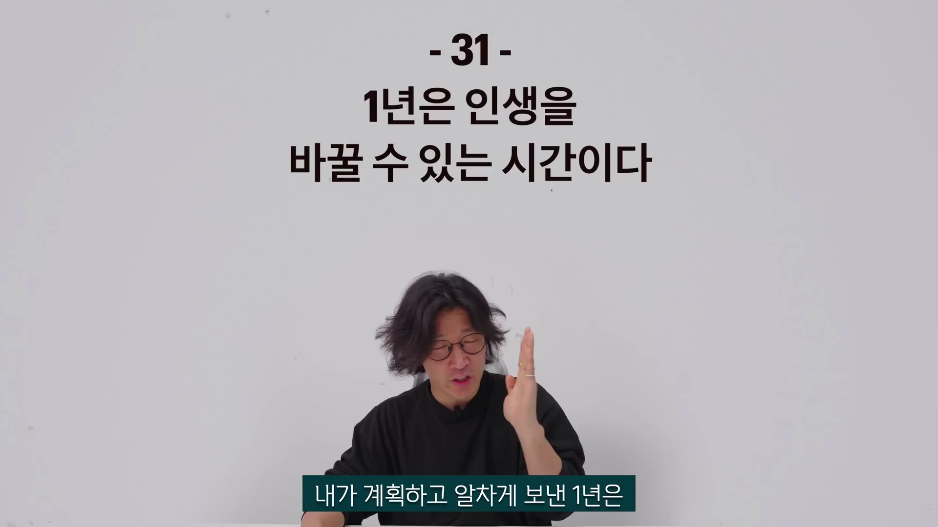 흰 배경 앞에 검은색 티셔츠를 입은 안경 쓴 남성이 손짓하며 이야기하고 있고, 머리 위에는 '1년은 인생을 바꿀 수 있는 시간이다'라는 문구가 적혀 있음.