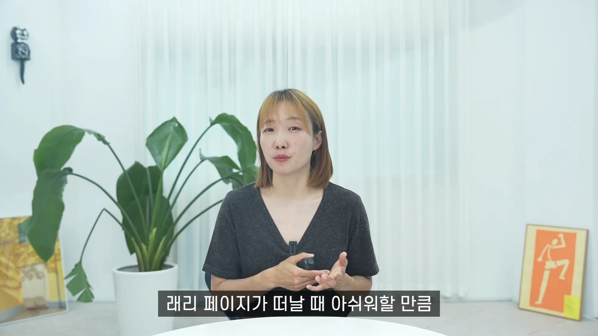 단발머리를 한 여성이 밝은 실내에서 시청자를 바라보며 손을 움직여 이야기하고 있고, 뒤쪽으로 대형 관엽식물과 그림 액자가 놓여 있습니다.