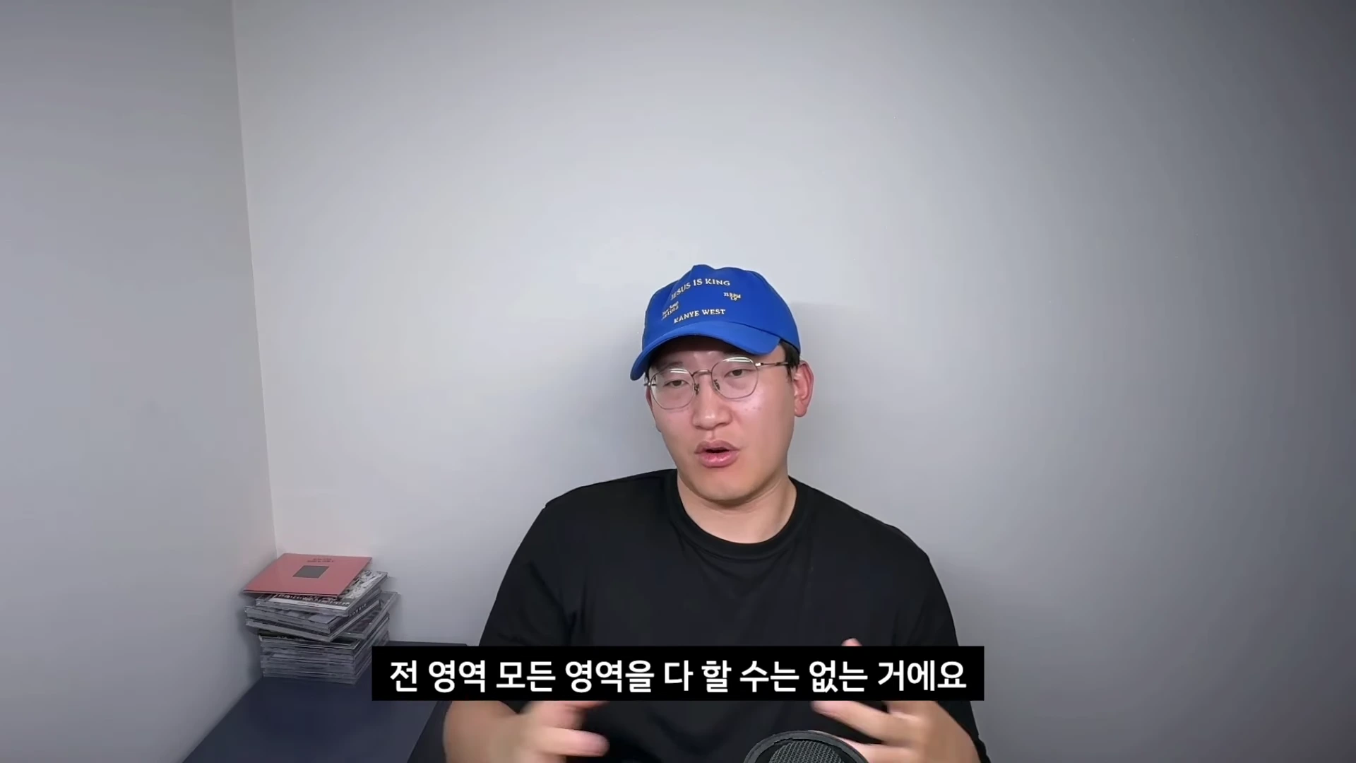 안경을 쓰고 파란색 볼캡을 쓴 남성이 흰 벽을 배경으로 앉아 손짓하며 이야기를 하고 있다. 화면 하단에는 자막이 떠 있다.